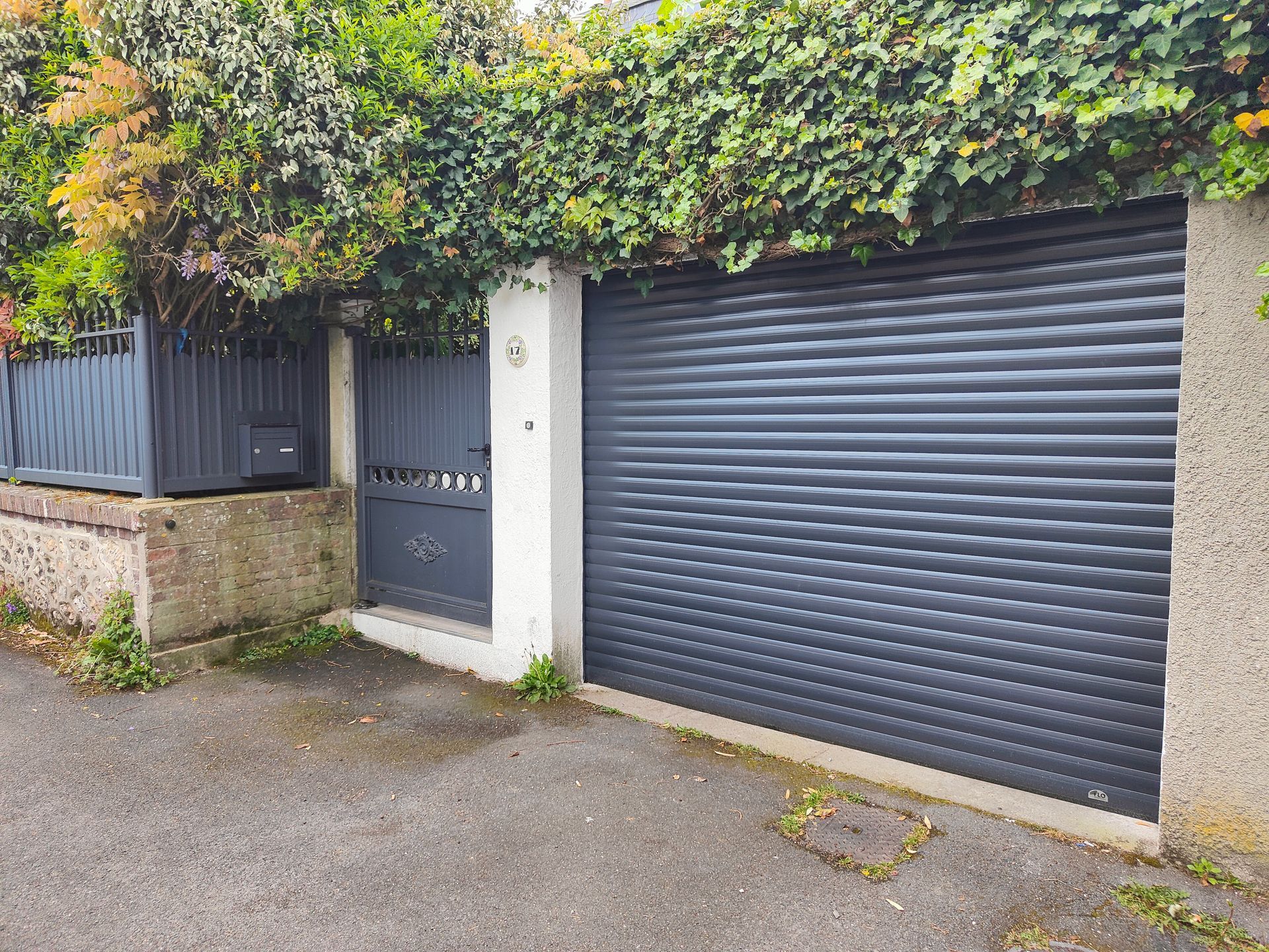Entrée de maison avec une porte de garage sombre, un portillon, un mur en pierre et une haie couverte de lierre au-dessus de l’allée