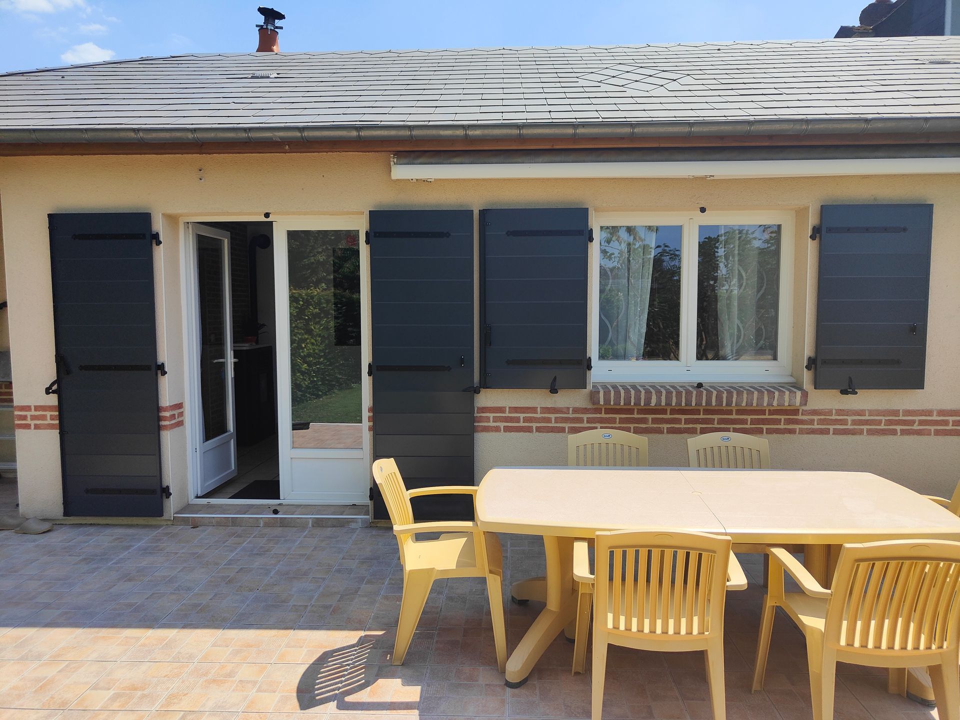Patio avec une façade de maison, une porte vitrée ouverte, des volets sombres et une table avec des chaises jaunes.