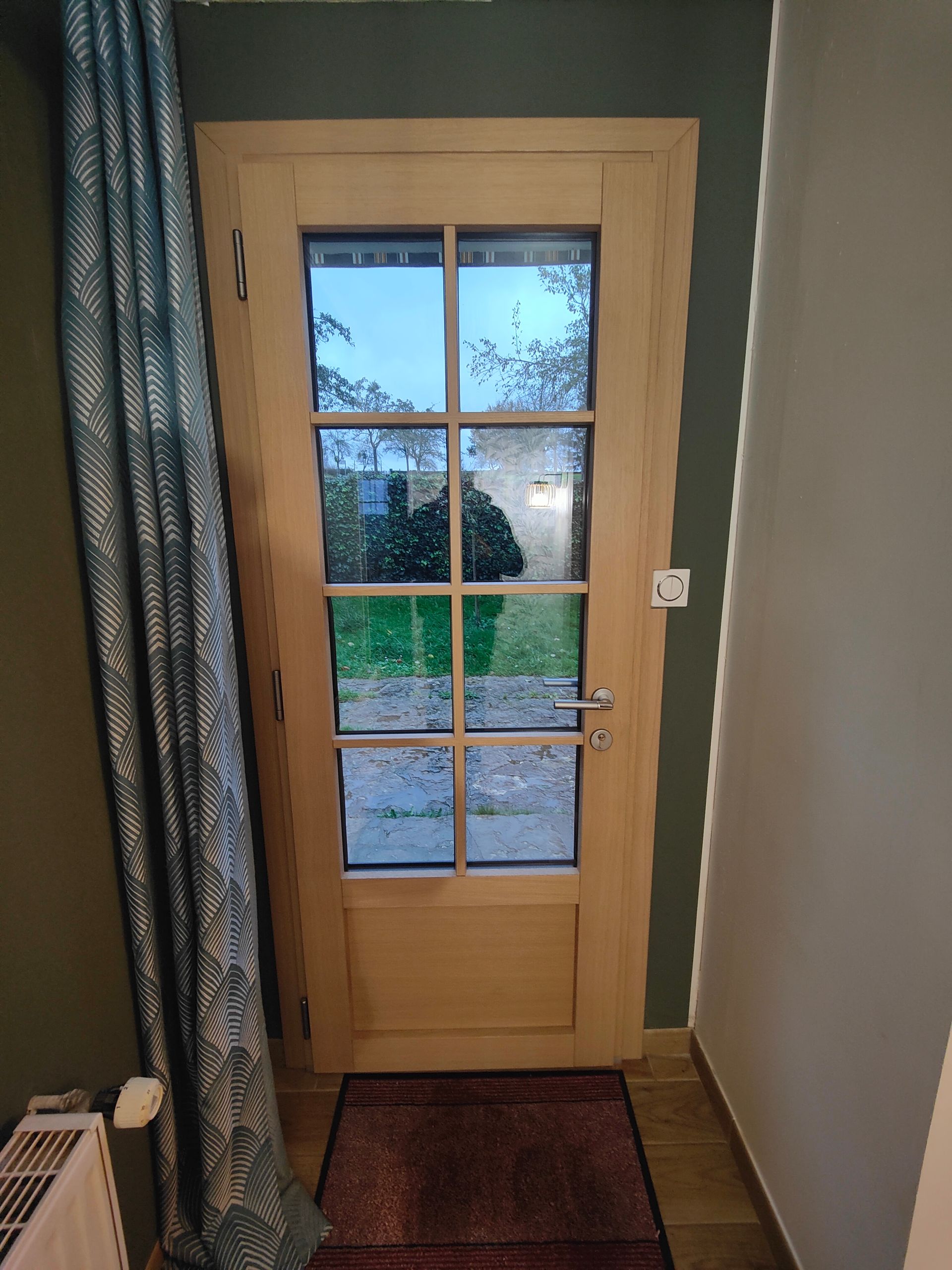 Porte d’entrée en bois avec des panneaux vitrés donnant sur une terrasse extérieure mouillée, avec un rideau à gauche et un interrupteur mural à proximité