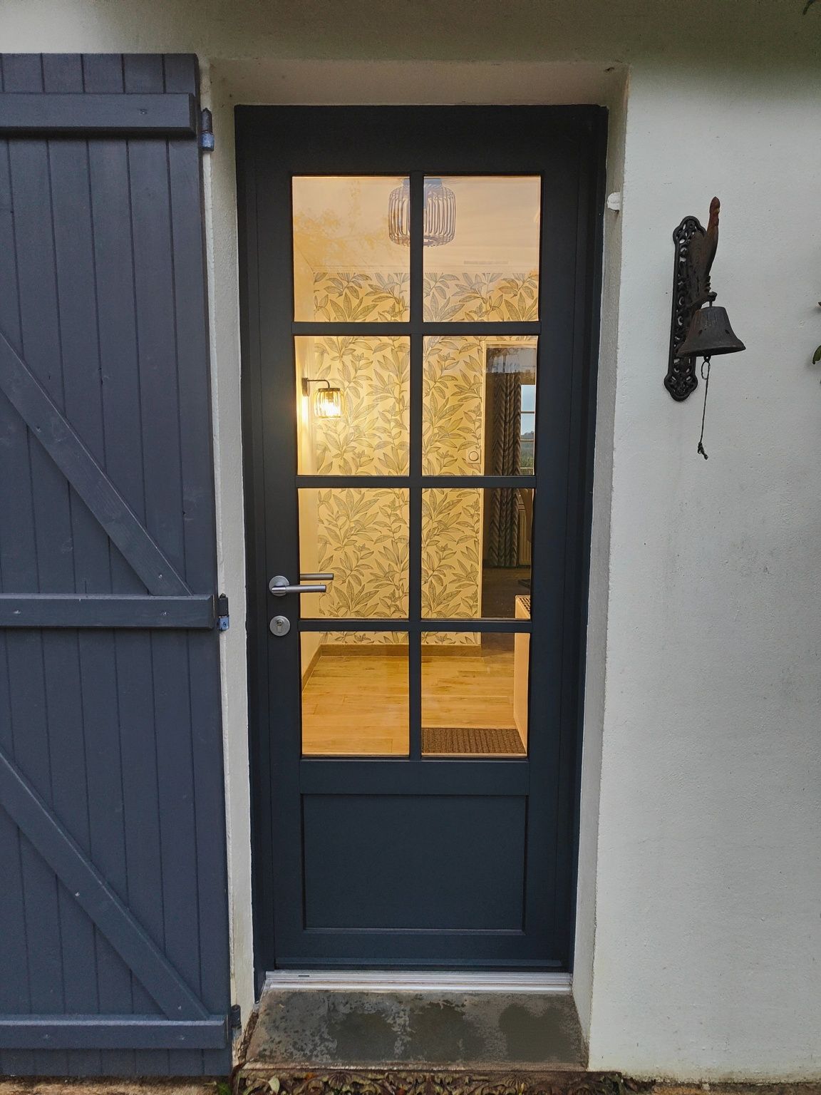Porte d’entrée bleu foncé vitrée, à côté d’un volet bleu et d’un mur blanc, éclairée de l’intérieur