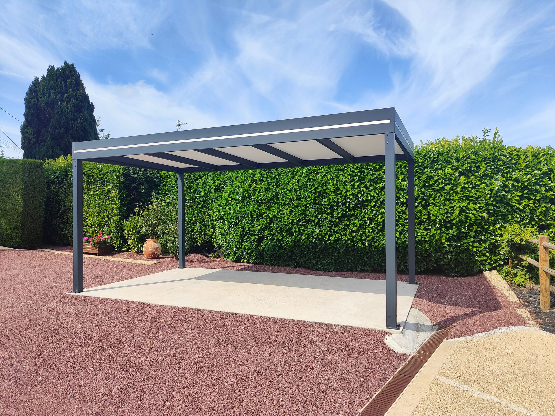 Pergola moderne grise sur une dalle de béton, près d’une haie verte et d’une cour en gravier rouge