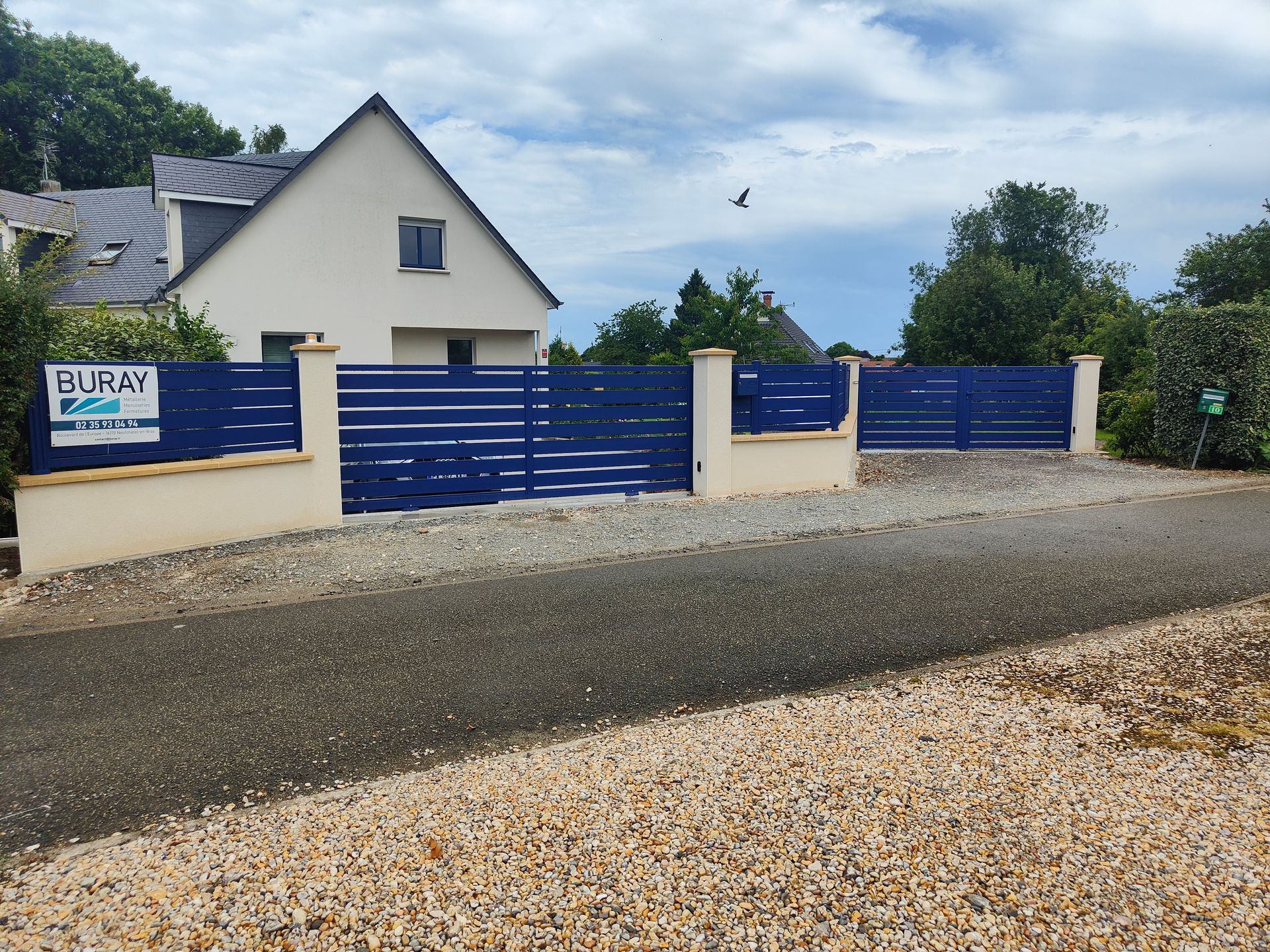 Portail et clôture bleus devant une maison blanche, au bord d’une route de gravier, sous un ciel nuageux