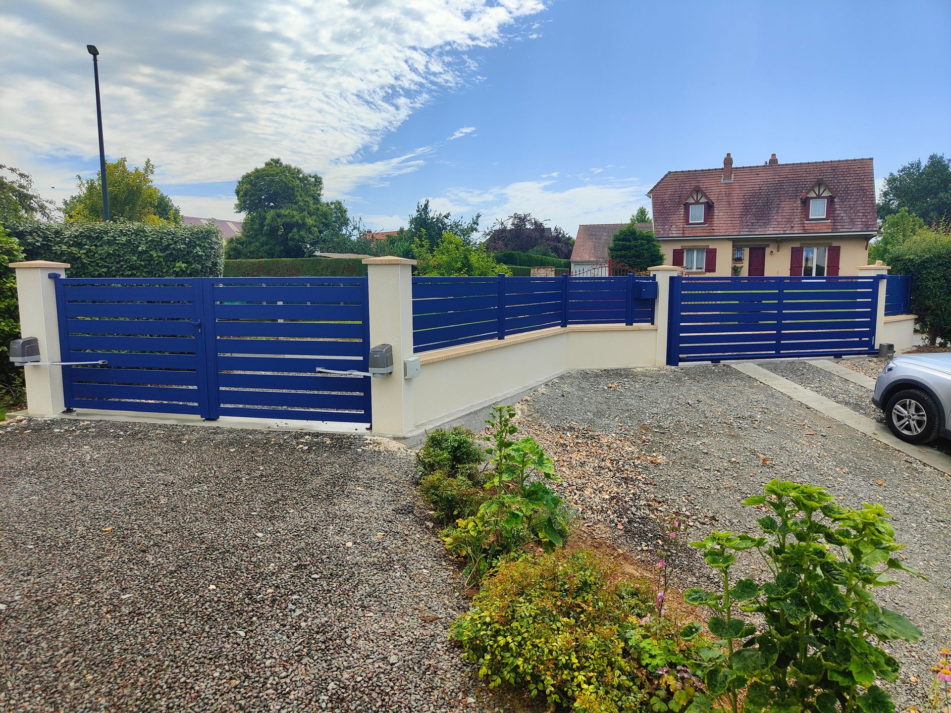 Portail d’entrée bleu avec des piliers crème, près de tas de gravier et d’une maison en arrière-plan