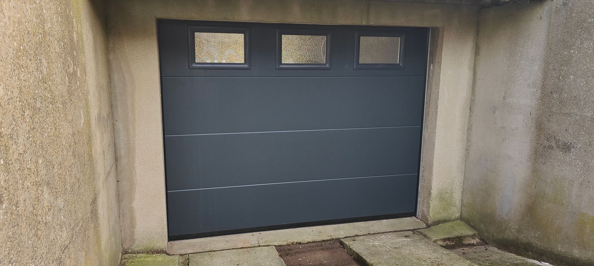Porte de garage gris foncé avec trois petites fenêtres, intégrée dans un mur en béton.