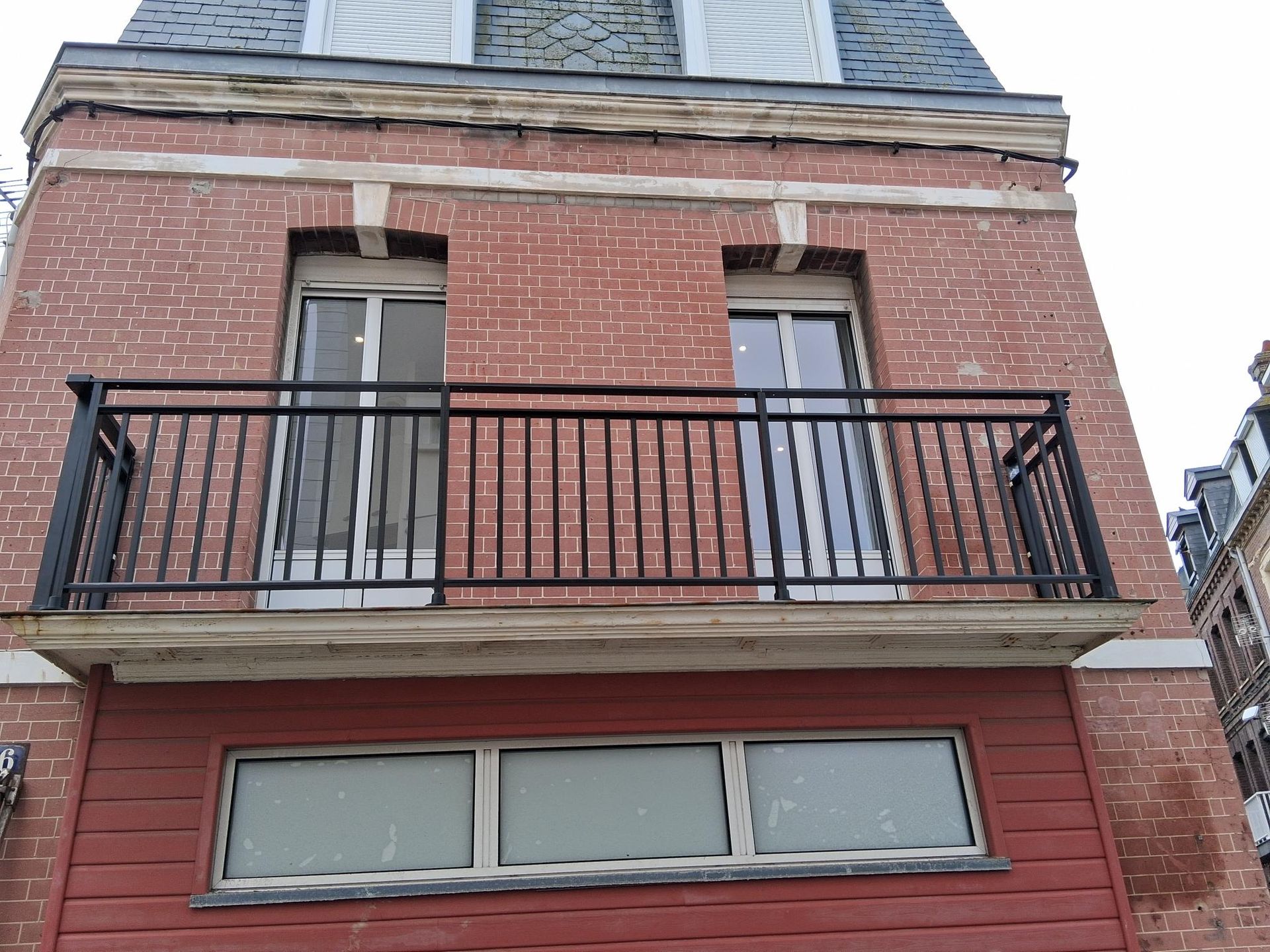 Façade d’un immeuble en briques avec un balcon noir et de hautes fenêtres au-dessus d’un rez-de-chaussée rouge