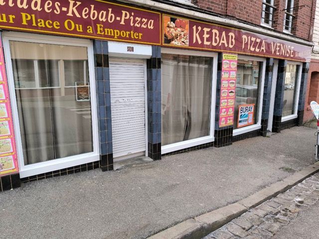 Vue de rue d’une devanture de vente à emporter de kebabs et de pizzas, avec une enseigne rouge et jaune et des volets baissés