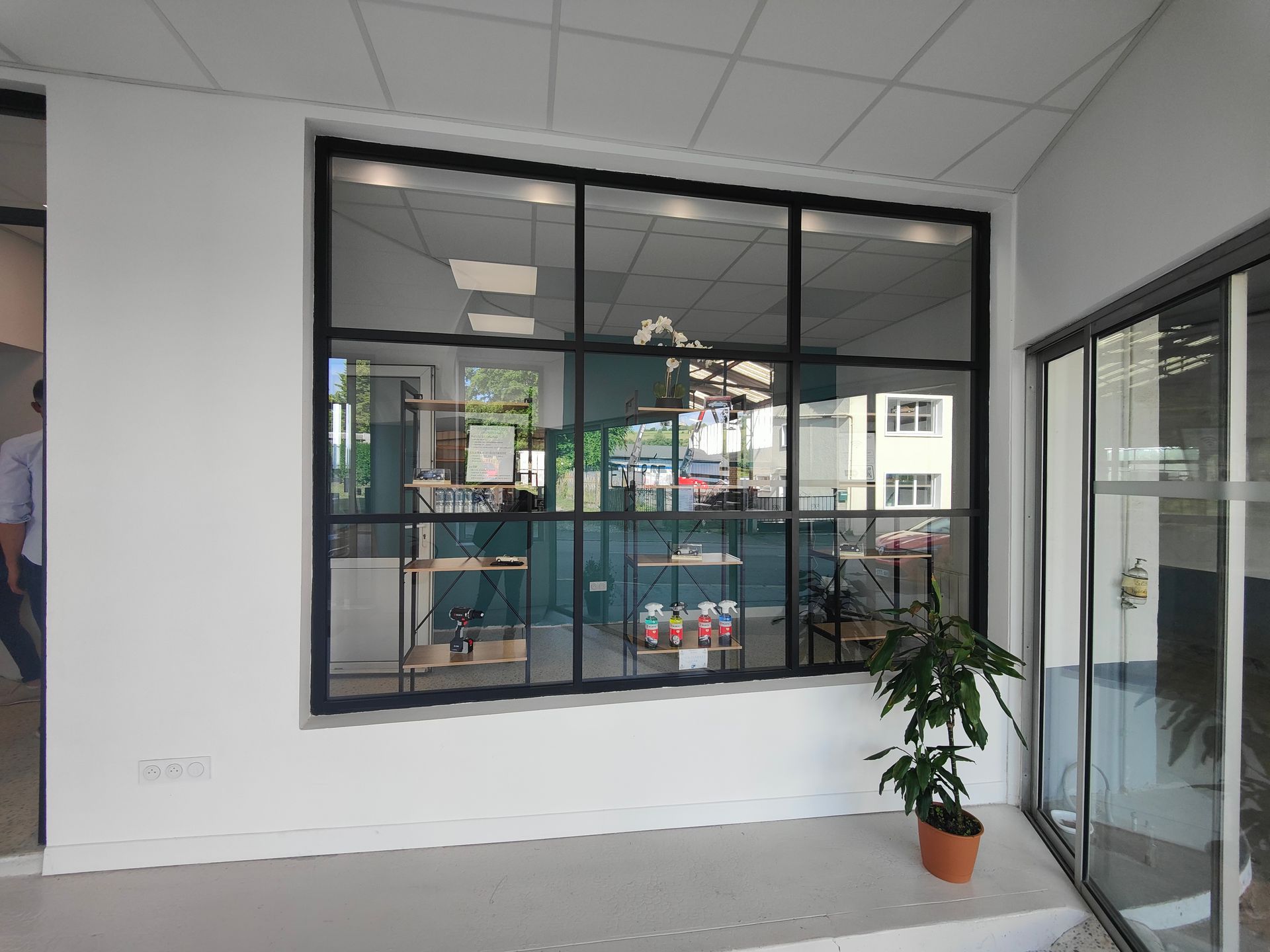 Intérieur de bureau avec une grande fenêtre à cadre noir, une porte vitrée et une plante en pot sur un sol carrelé.