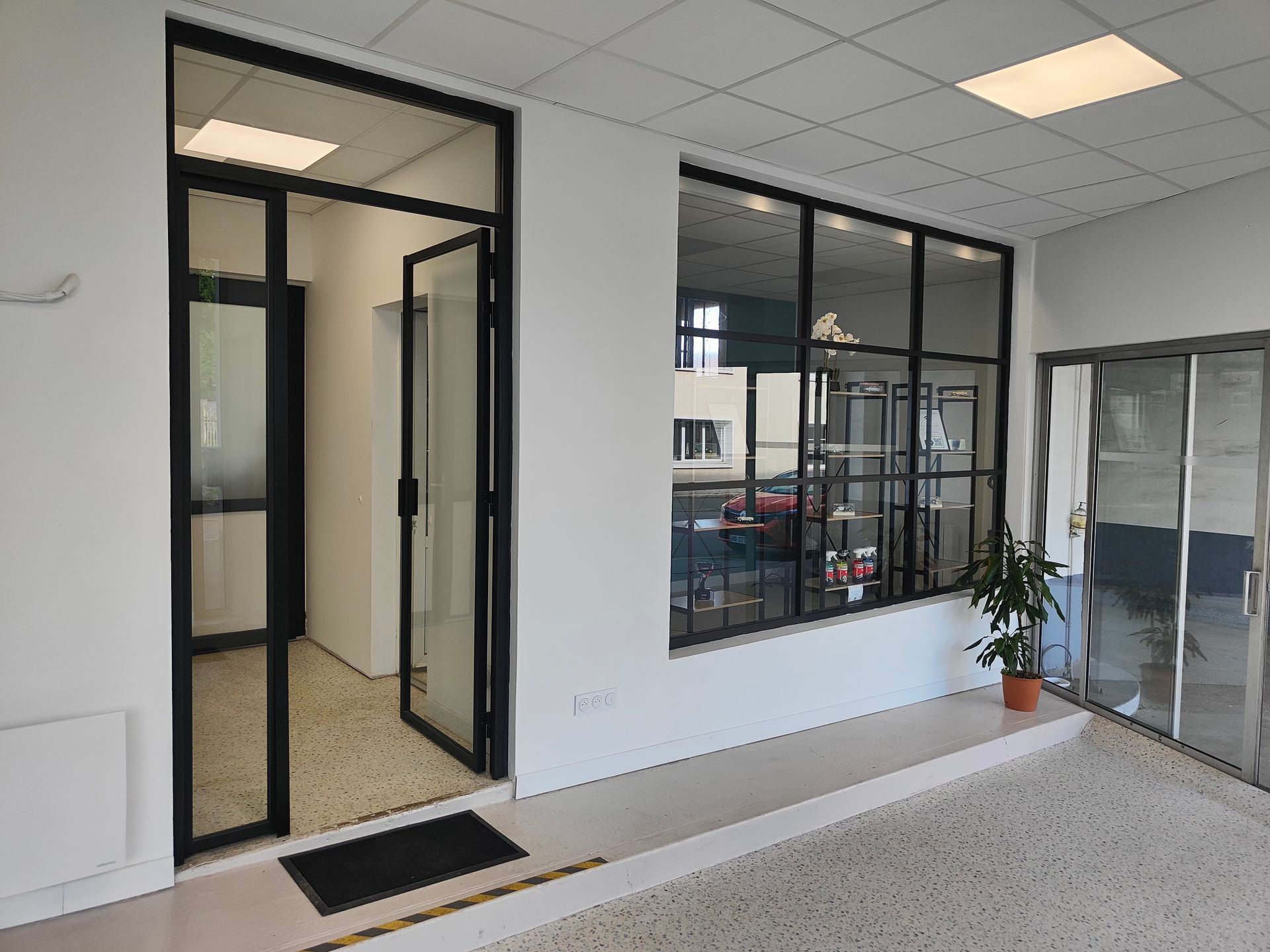 Hall d’accueil de bureaux avec des portes vitrées, de grandes fenêtres et une plante en pot près de l’entrée.