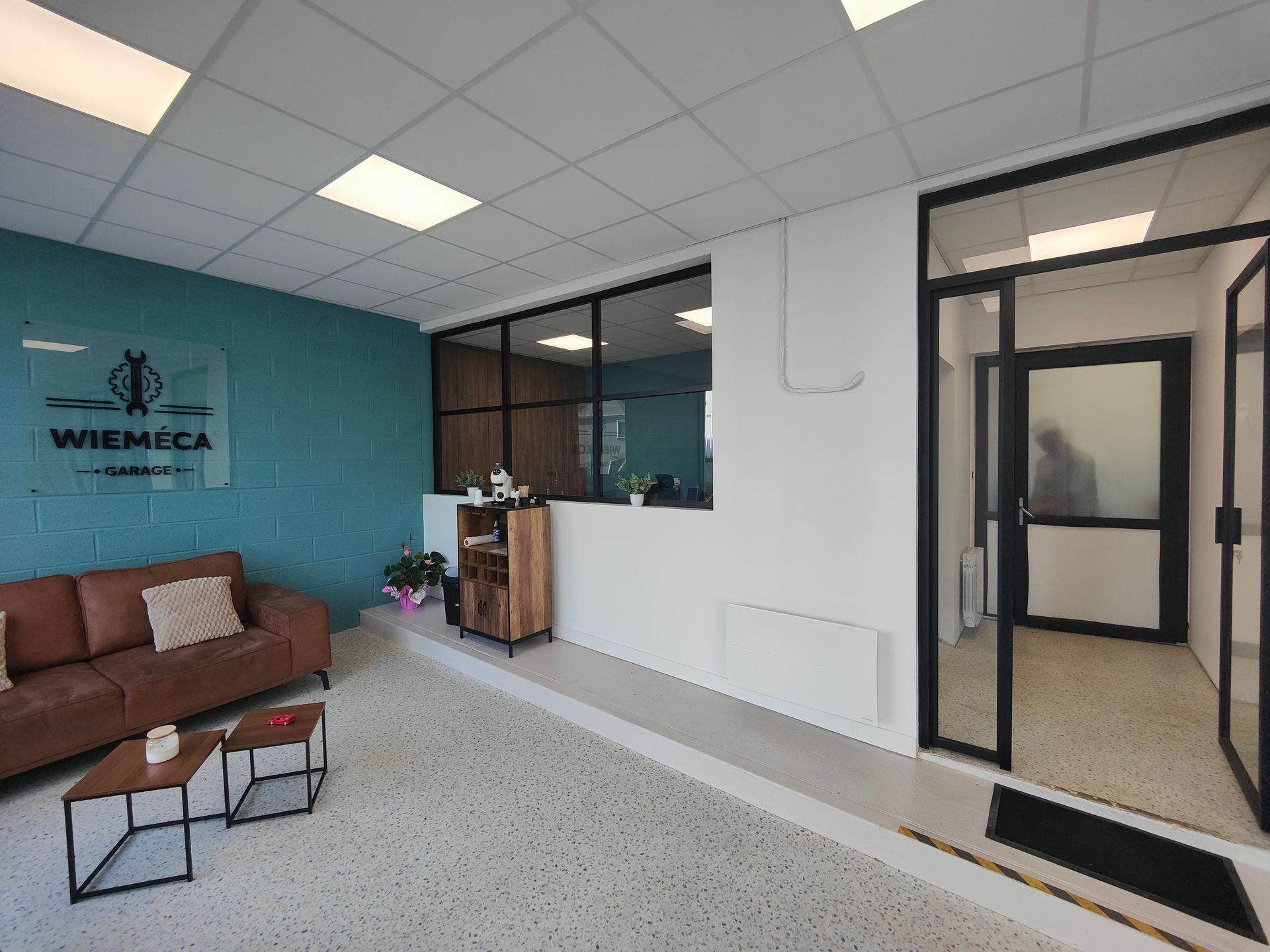 Hall d’entrée moderne de bureaux avec un mur d’accent bleu canard, un canapé marron, des pièces aux cloisons vitrées et un espace d’accueil.