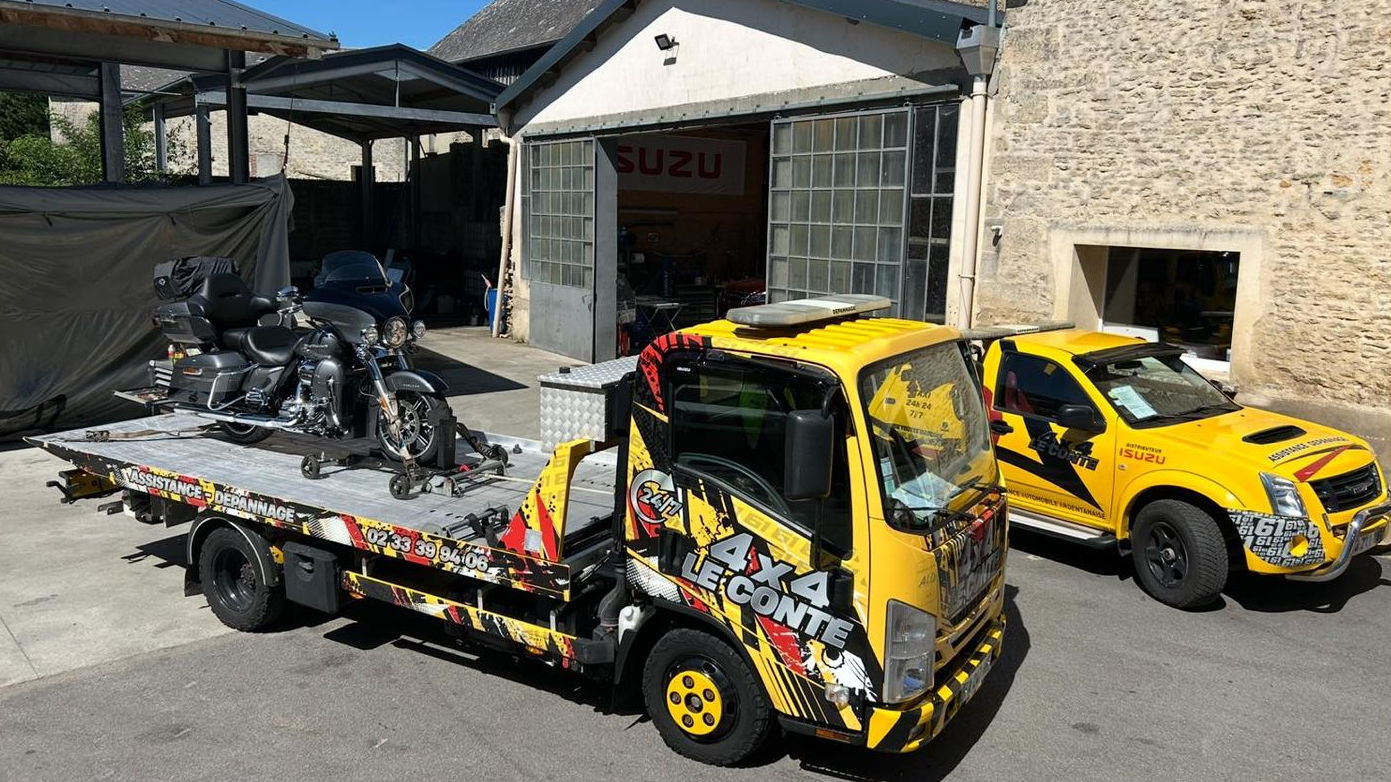 Dépanneuse jaune et voiture avec graphismes garés devant un garage, avec des motos sur la plateforme du camion.