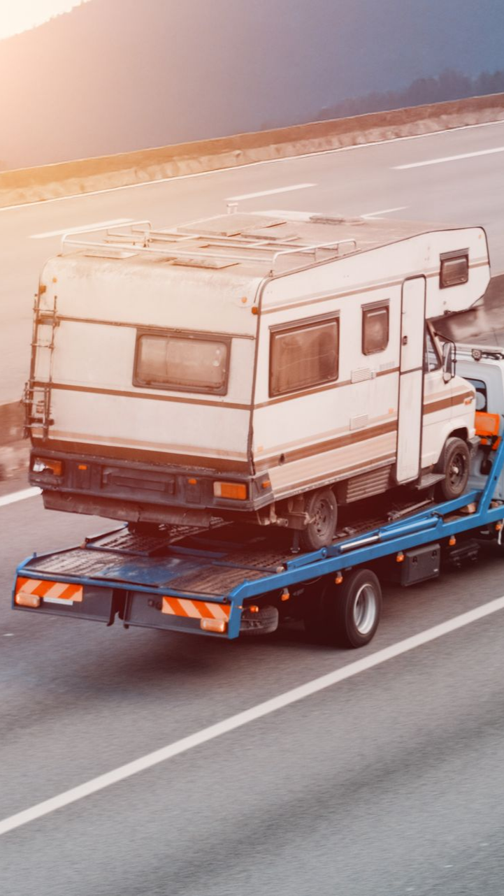 Un camping-car remorqué sur un camion plateau le long d'une autoroute. Le soleil brille, mettant en valeur les véhicules.