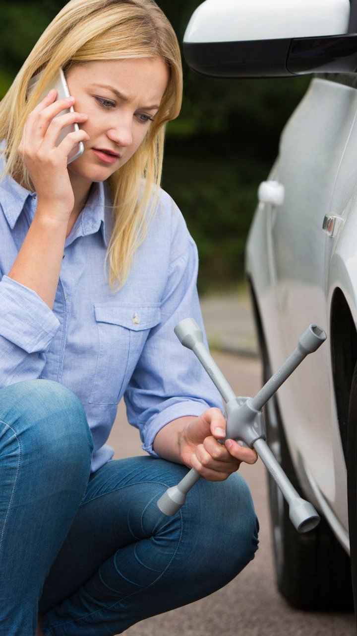 Femme au téléphone, accroupie près d'une voiture avec une clé à molette, l'air inquiète.