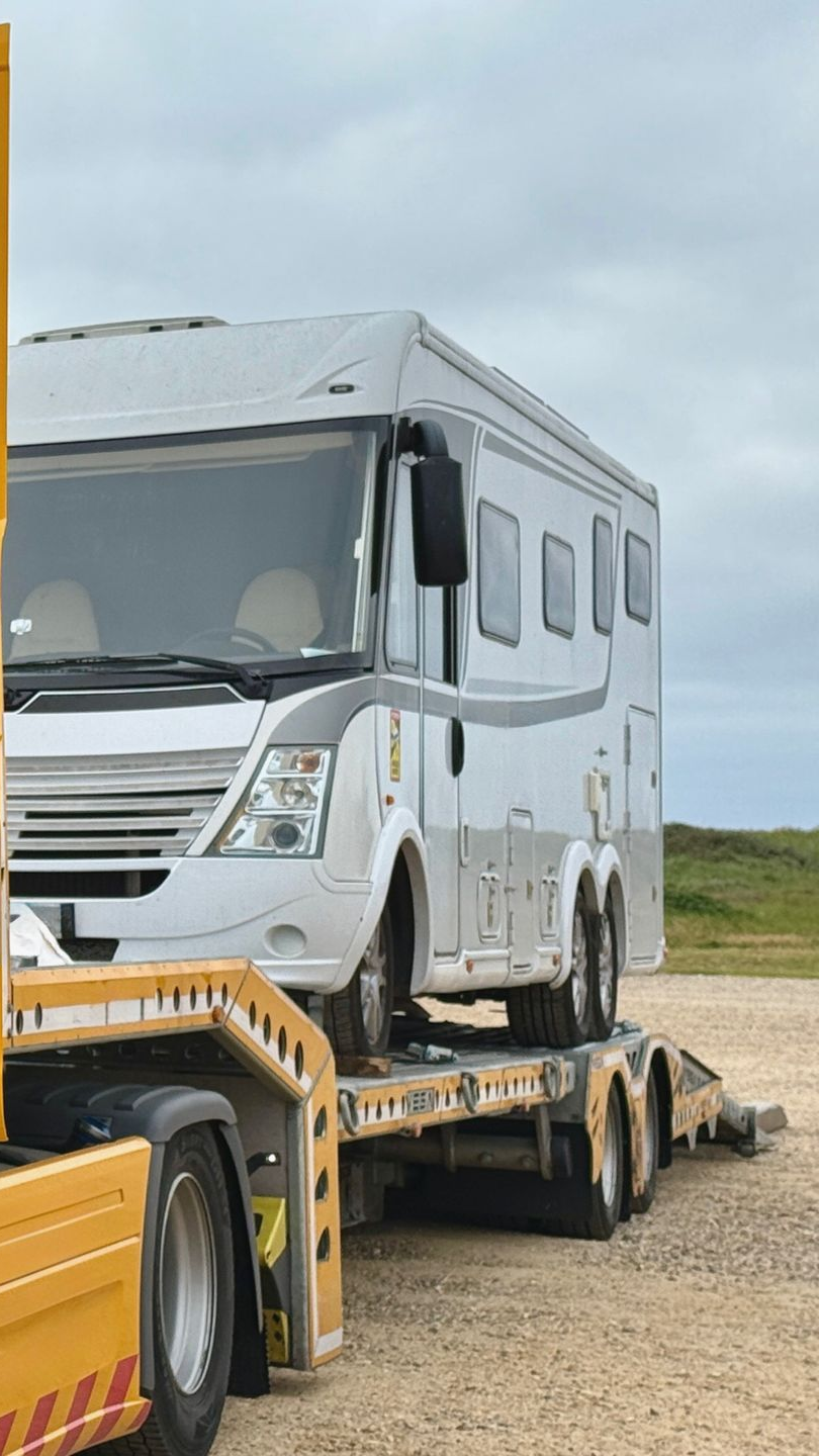 Camping-car transporté sur un grand camion à plateau, sur une route de gravier sous un ciel nuageux.