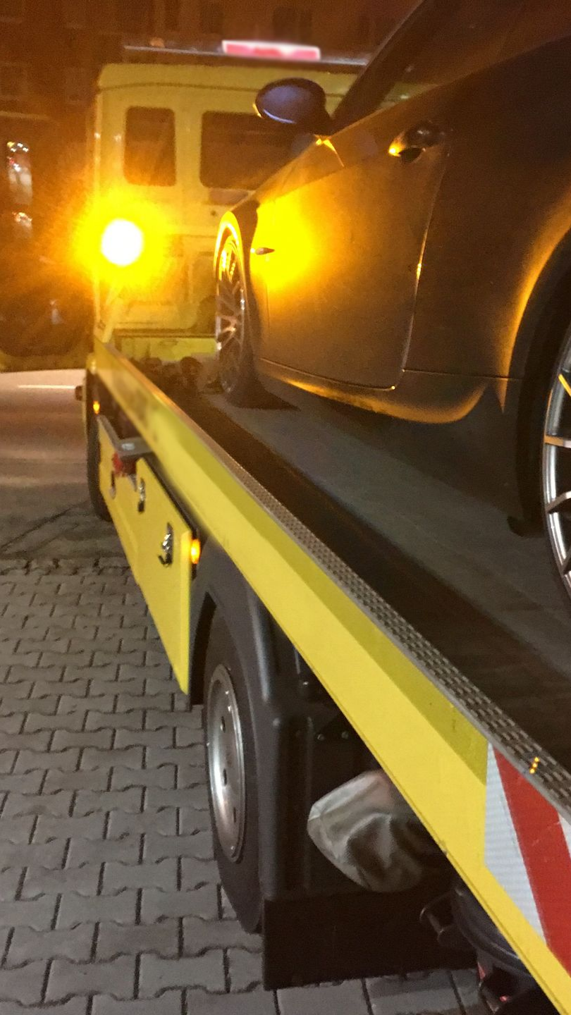 Une voiture sombre remorquée sur une dépanneuse à plateau la nuit, éclairée par un feu de détresse orange.