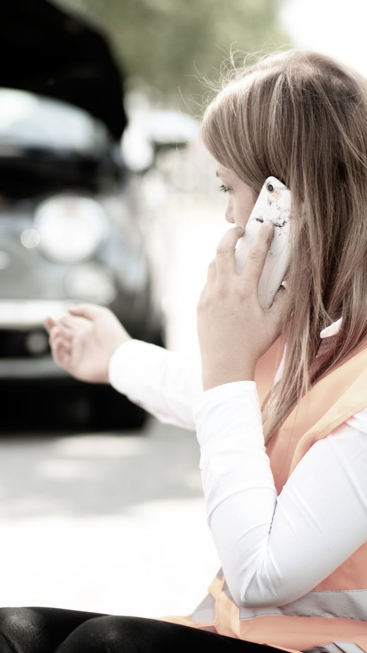 Une femme au téléphone près d'une voiture, capot relevé, probablement en panne. Elle porte un gilet de sécurité.