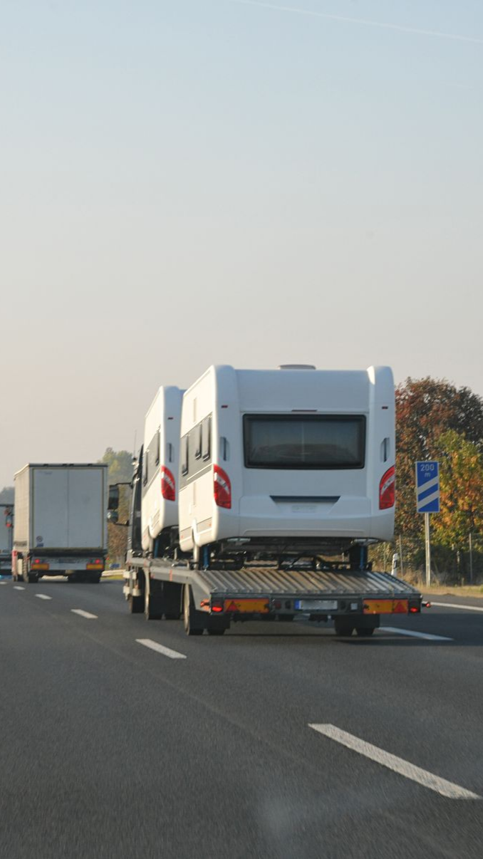 Deux caravanes blanches sur un camion à plateau sur une autoroute ; d'autres véhicules en arrière-plan.