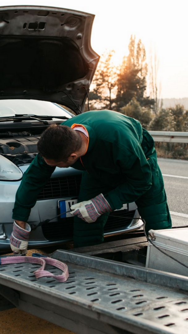 Opérateur de dépanneuse sécurisant une voiture avec le capot ouvert, sur le bord de la route au coucher du soleil.