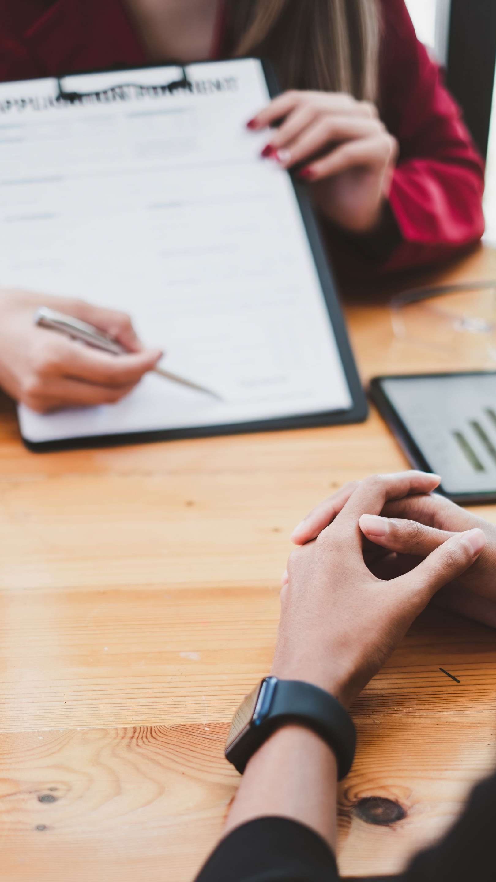 Une personne désigne un document avec un stylo lors d'une réunion ; une autre personne joint les mains. Table en bois, bureau.