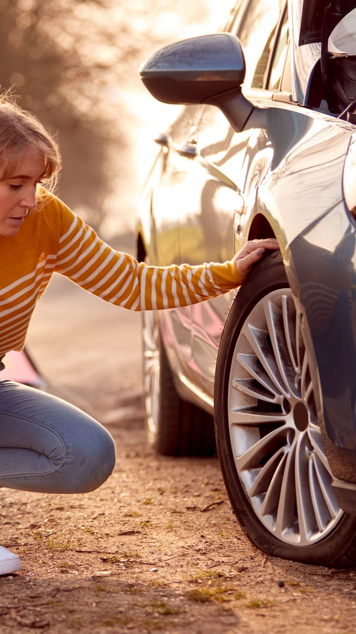 Une femme vérifie le pneu crevé d'une voiture sur une route rurale, l'air inquiète, avec la lumière du soleil et une voiture bleue.