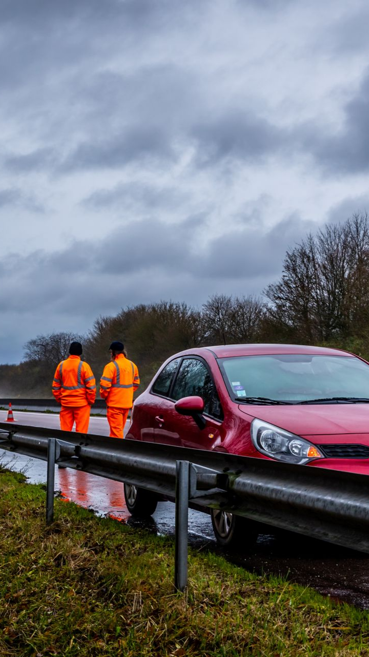 Une voiture rouge coincée sur une glissière de sécurité avec deux ouvriers en gilet orange debout à proximité sur le bord d'une route inondée.