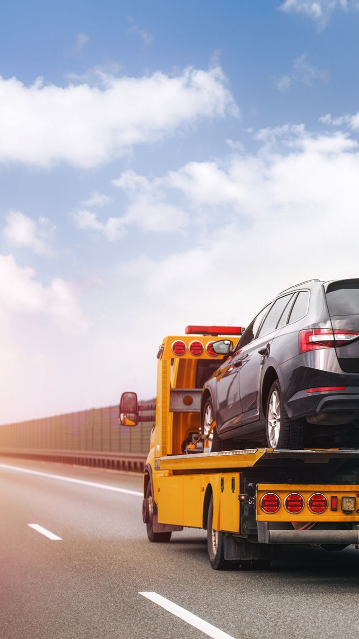 Une voiture noire sur une dépanneuse jaune roulant sur une autoroute sous un ciel bleu et nuageux.