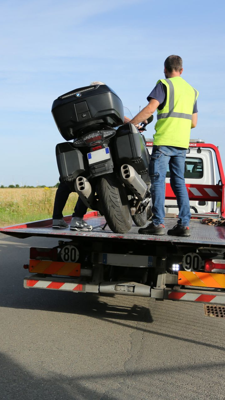 Un homme portant un gilet de sécurité charge une moto sur une dépanneuse sur une route rurale.