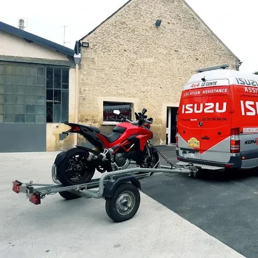 Moto rouge sur une remorque attelée à un fourgon d'assistance Isuzu, devant un immeuble.
