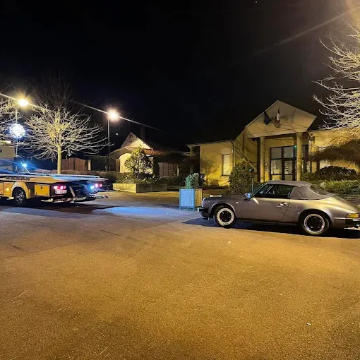 Une voiture classique chargée sur une remorque la nuit devant un bâtiment éclairé.