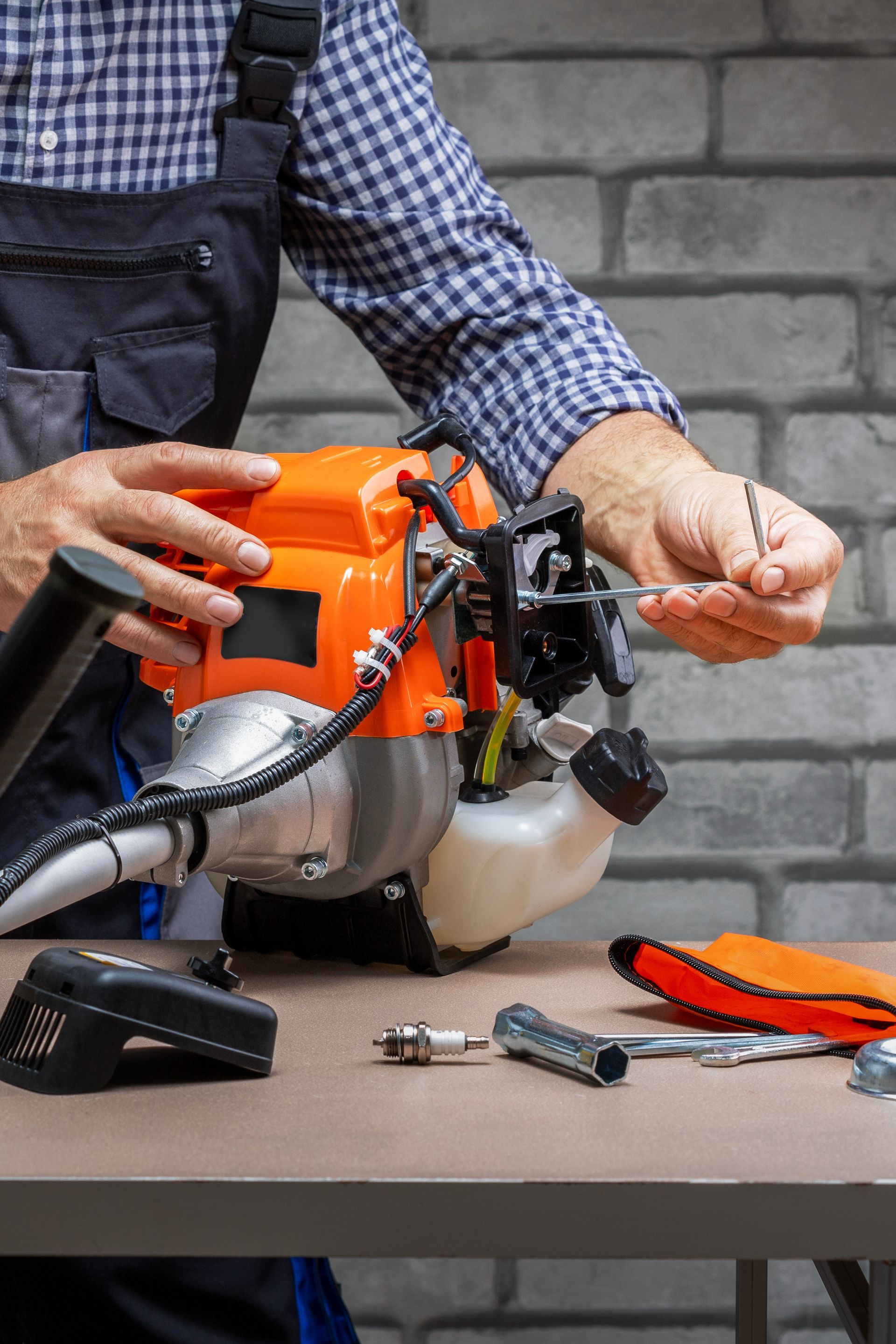 Un homme en salopette répare un coupe-herbe orange sur une table. Des outils sont à proximité, avec un mur de briques grises en arrière-plan.