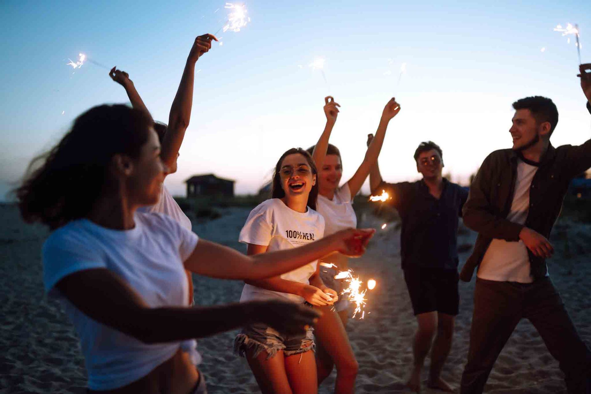 Amigos en una playa al anochecer sosteniendo bengalas, sonriendo y bailando.