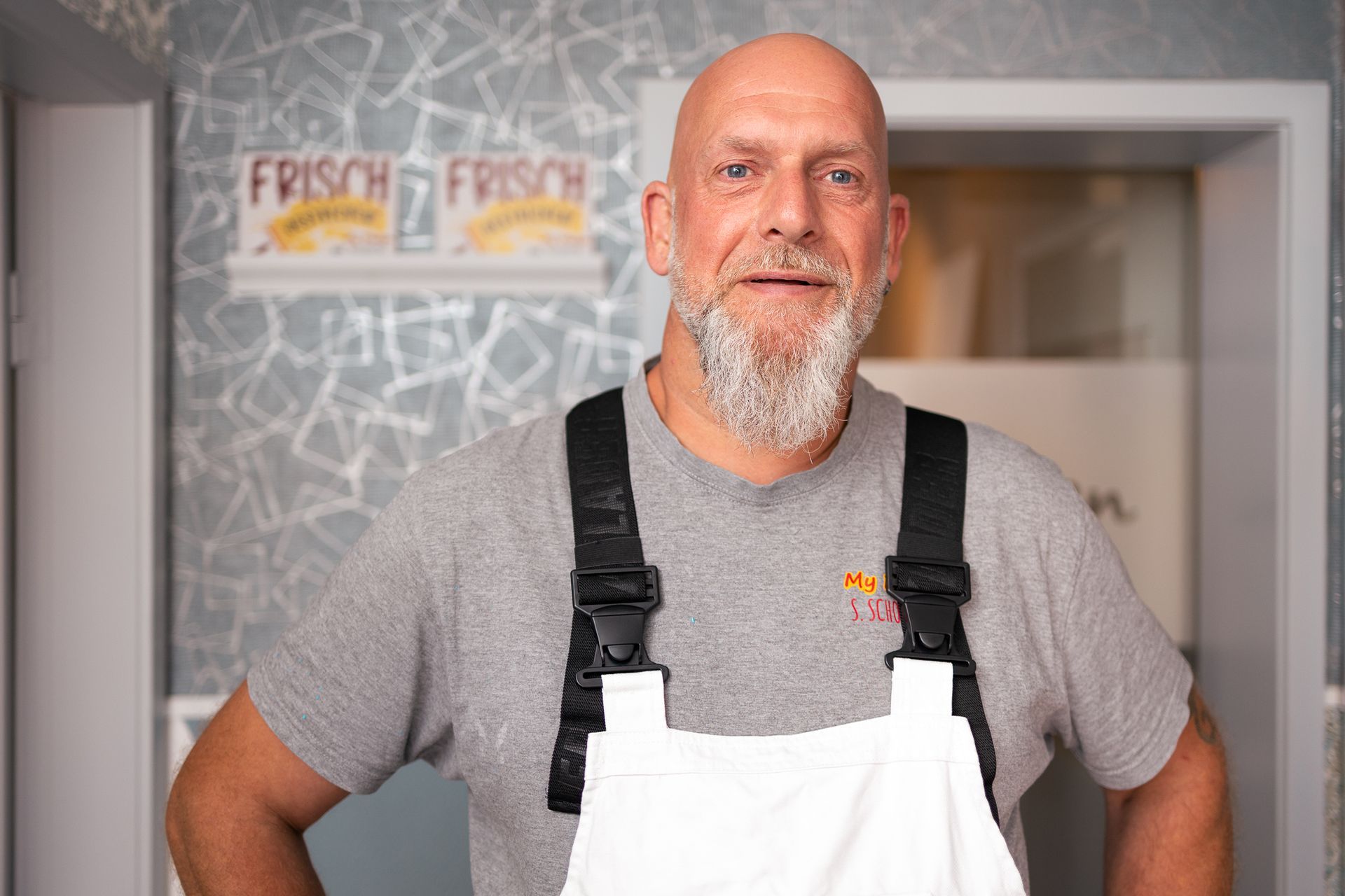 Ein kahlköpfiger Mann mit Bart trägt einen weißen Overall und ein graues Hemd.