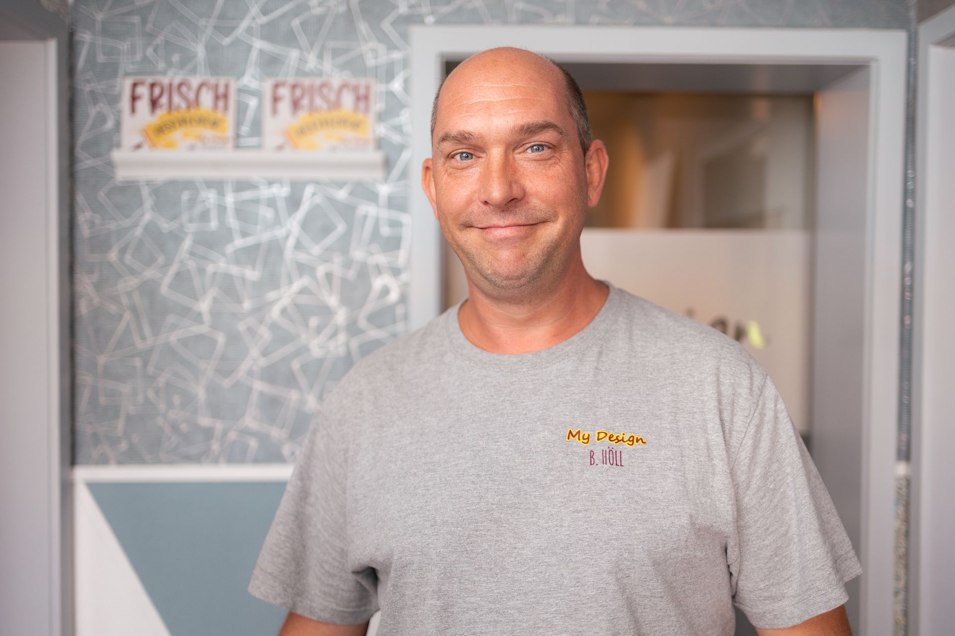 Ein kahlköpfiger Mann mit einem grauen T-Shirt steht vor einer Tür.