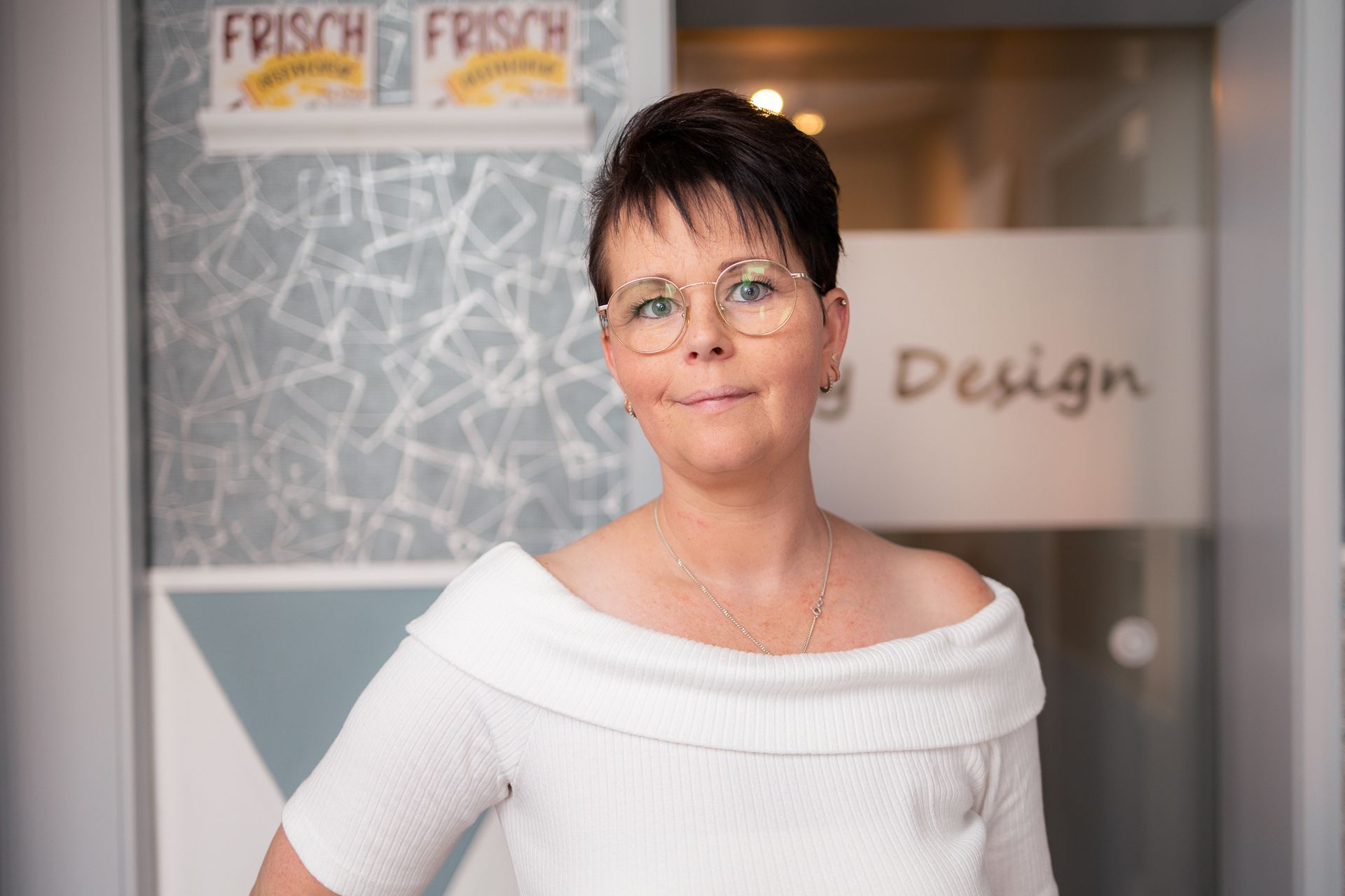 Eine Frau mit Brille und weißem Oberteil steht vor einer Glastür.
