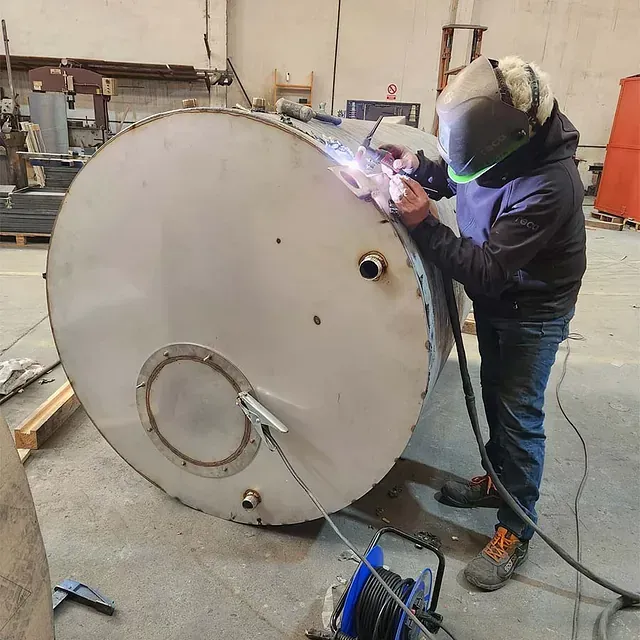 Un hombre está soldando un gran tanque de acero inoxidable.