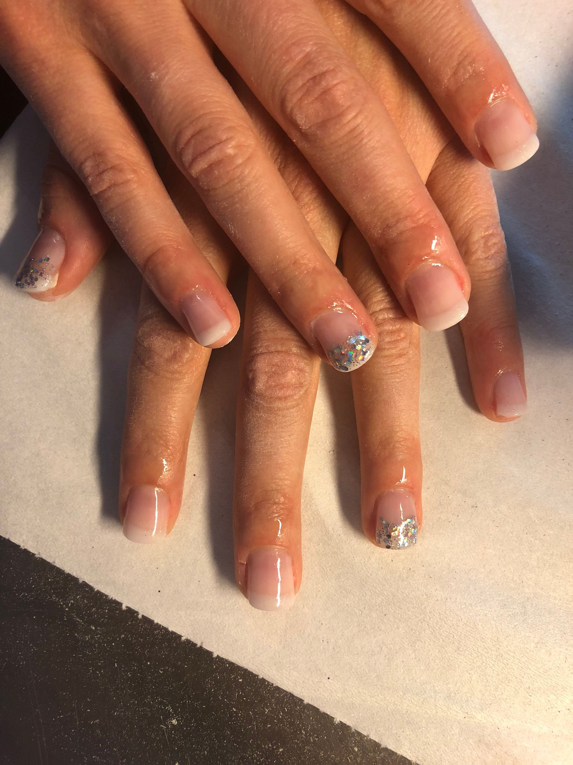 Des mains aux ongles naturels et aux accents de paillettes argentées reposent sur une surface claire.