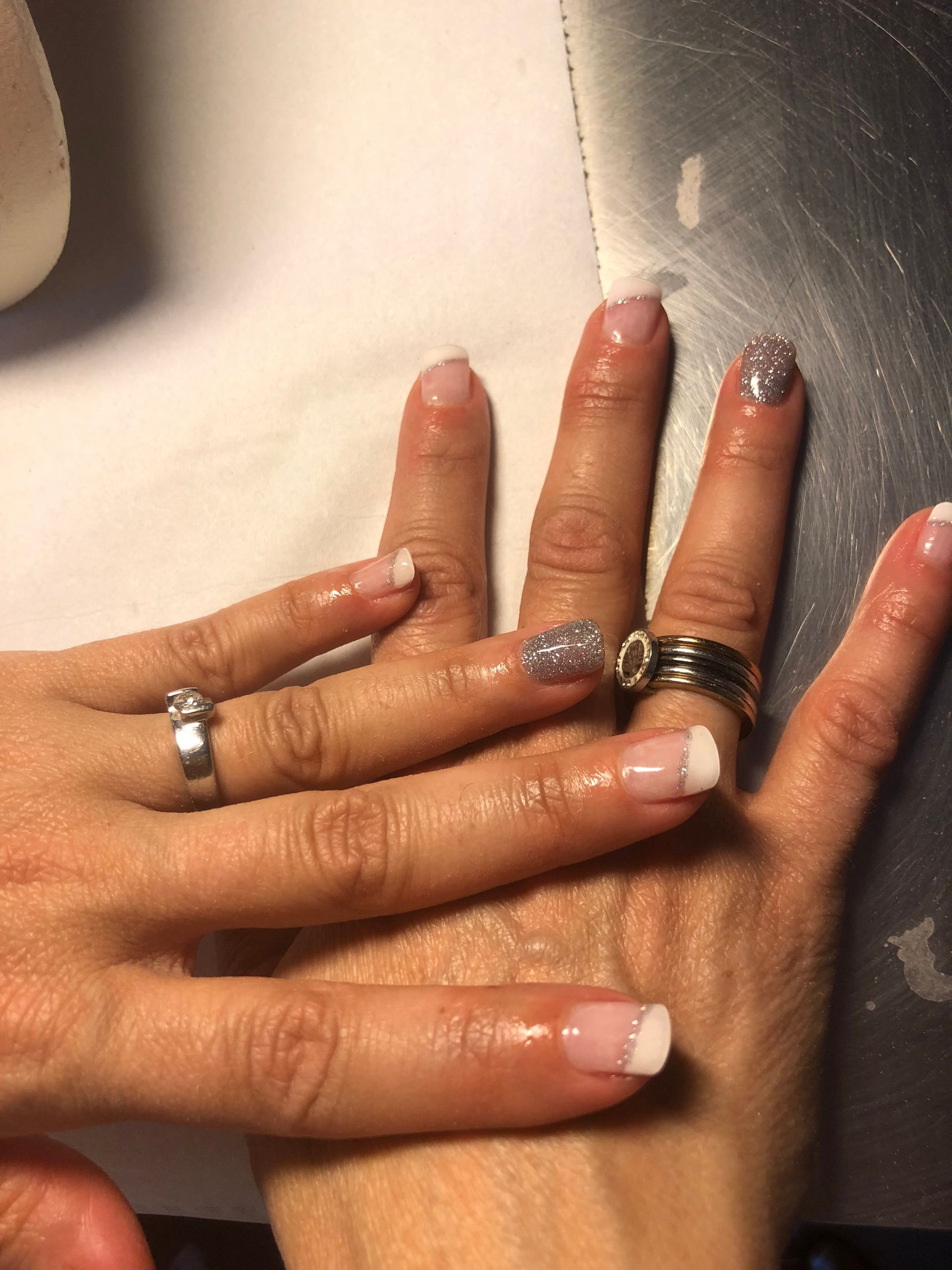 Quatre mains jointes, dévoilant des bagues en argent et un vernis à ongles à paillettes bleu-gris.