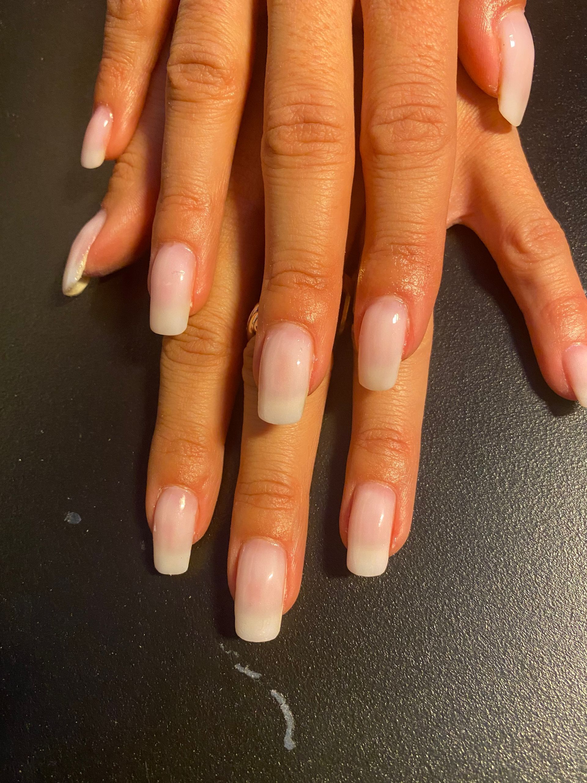 Des mains aux longs ongles nude ombrés, manucurés, reposent sur une surface sombre.