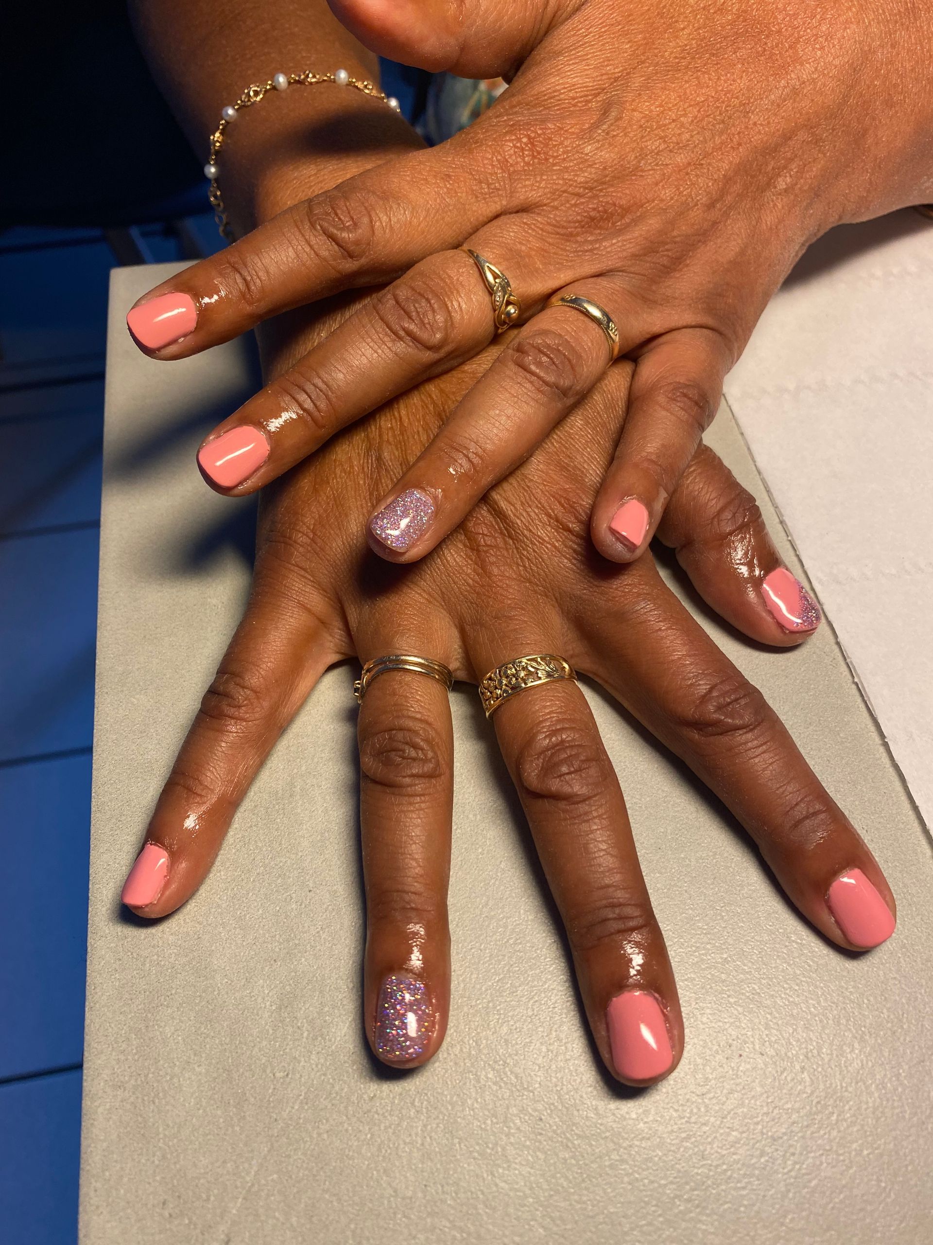 Des mains aux ongles vernis rose et lavande, ornées de bagues en or, sont posées côte à côte sur une table.
