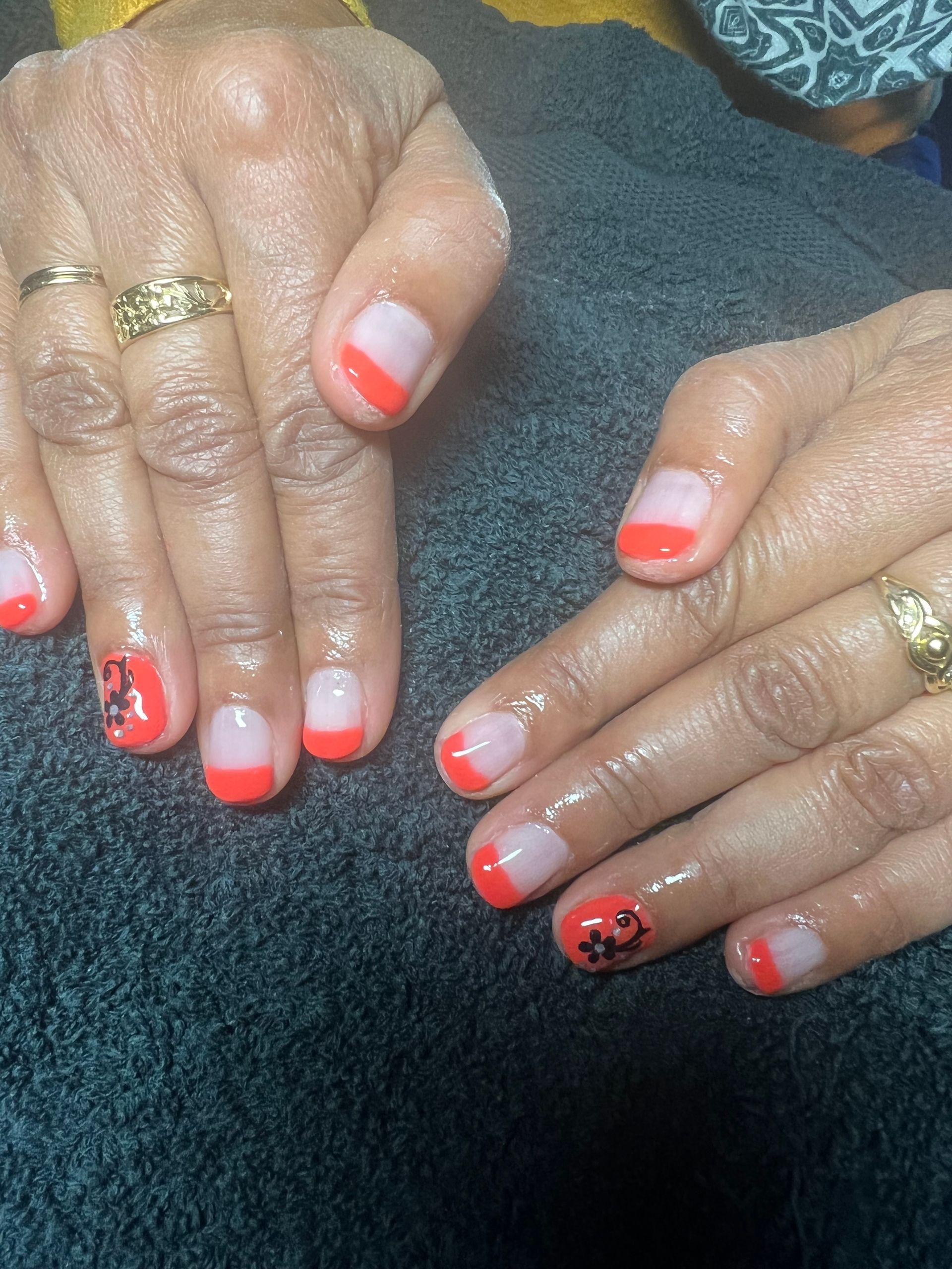 Des mains aux ongles corail vernis à la française et ornées de bagues en or reposent sur une serviette sombre.