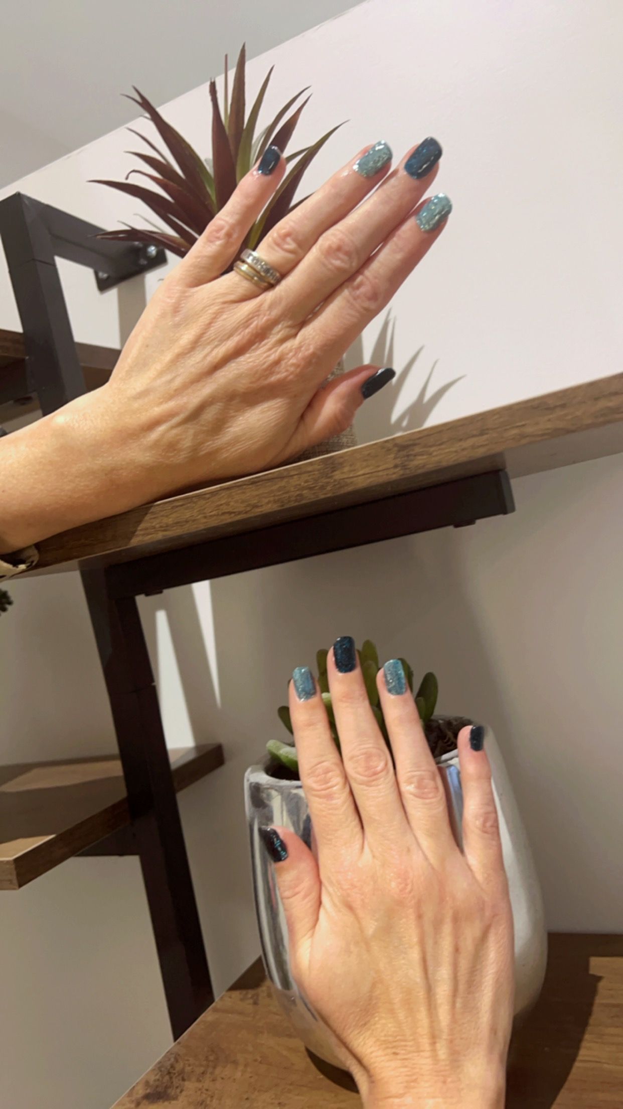 Deux mains aux ongles bleu-vert, l'une reflétée dans un miroir à côté d'une petite plante.