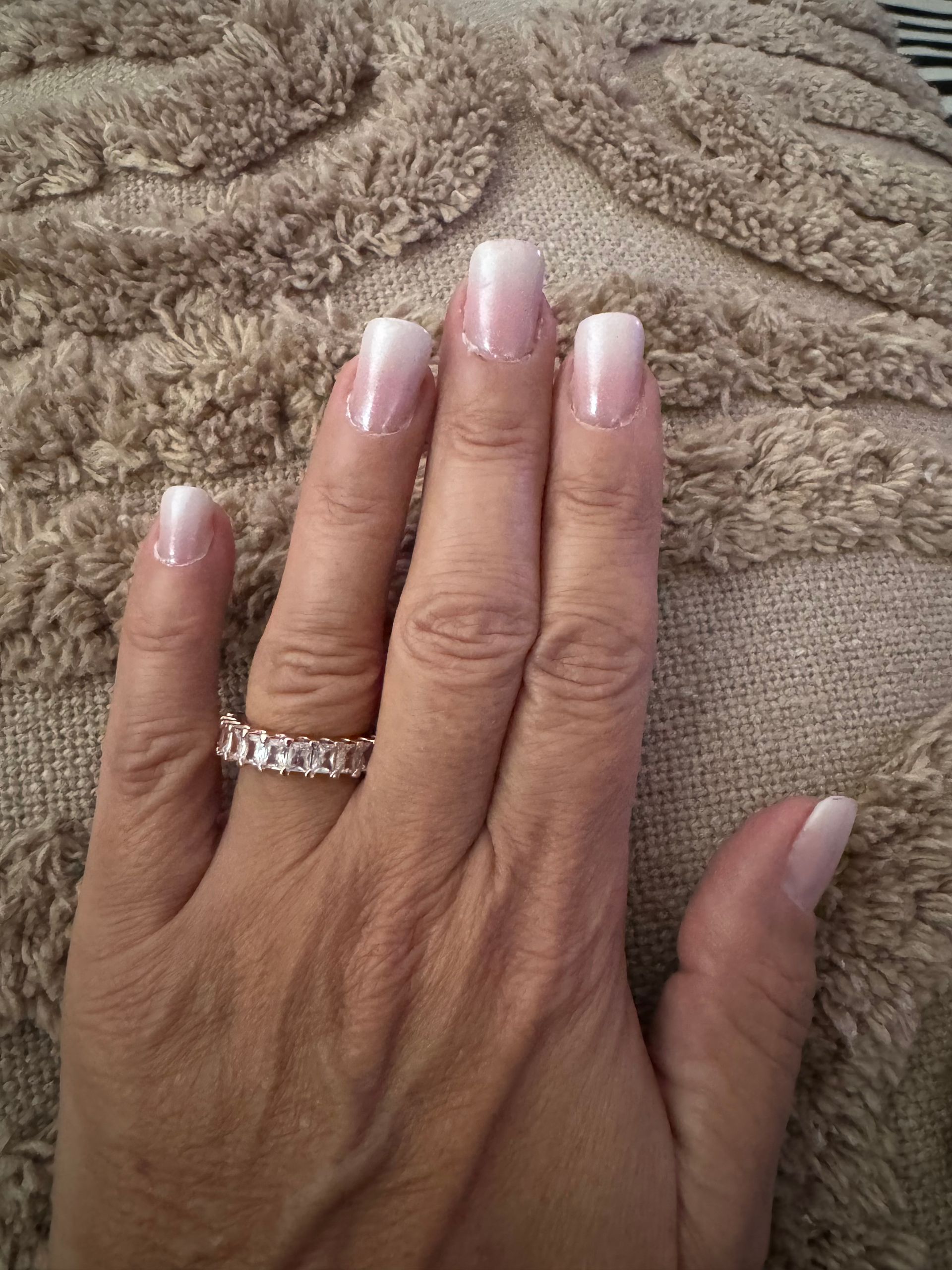 Main ornée d'une manucure française et d'une bague en diamants, posée sur une couverture beige texturée.
