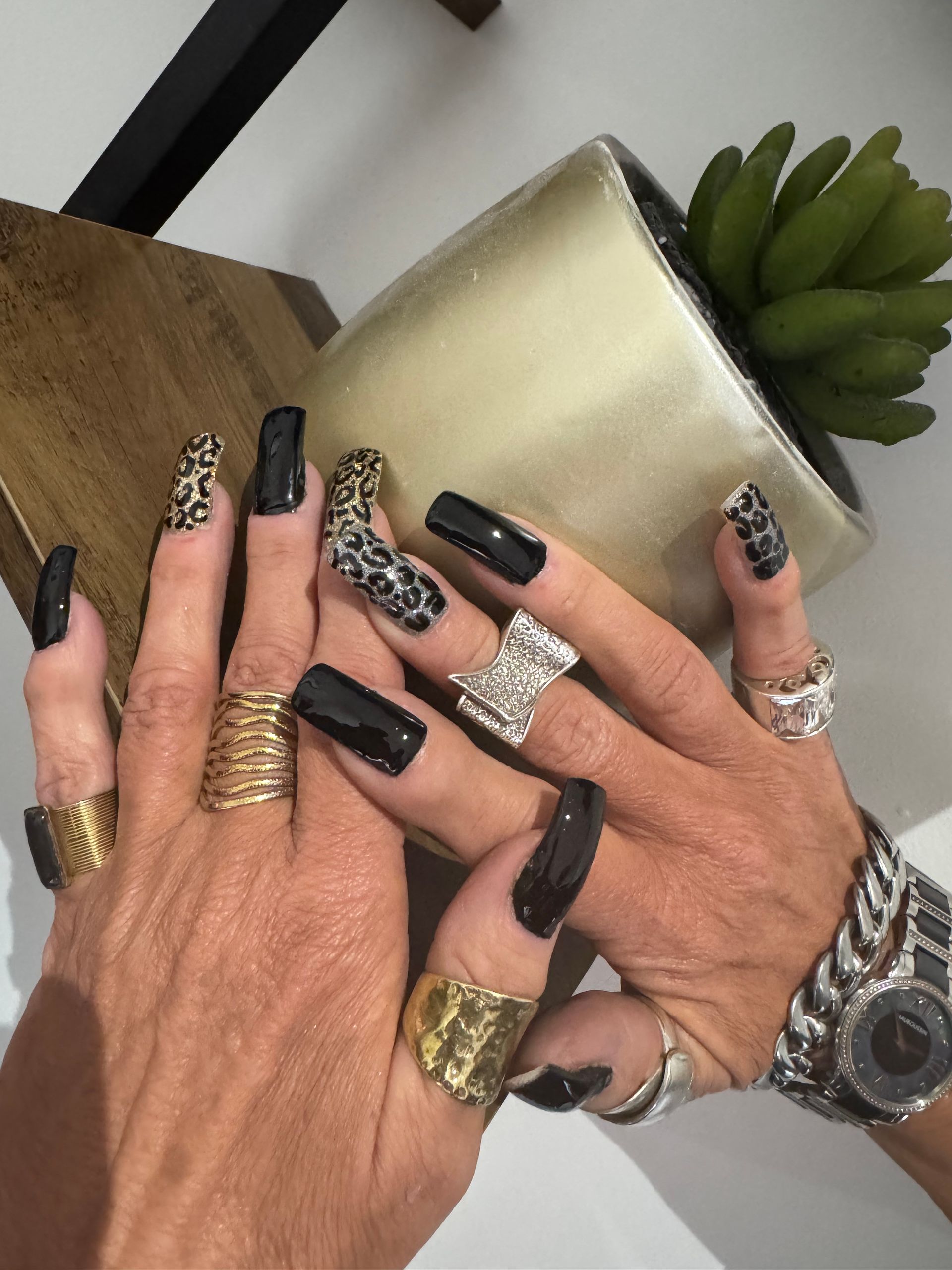 Gros plan sur des mains aux longs ongles noirs à motifs, portant des bagues empilées et une montre, près d'une plante et d'une bougie.