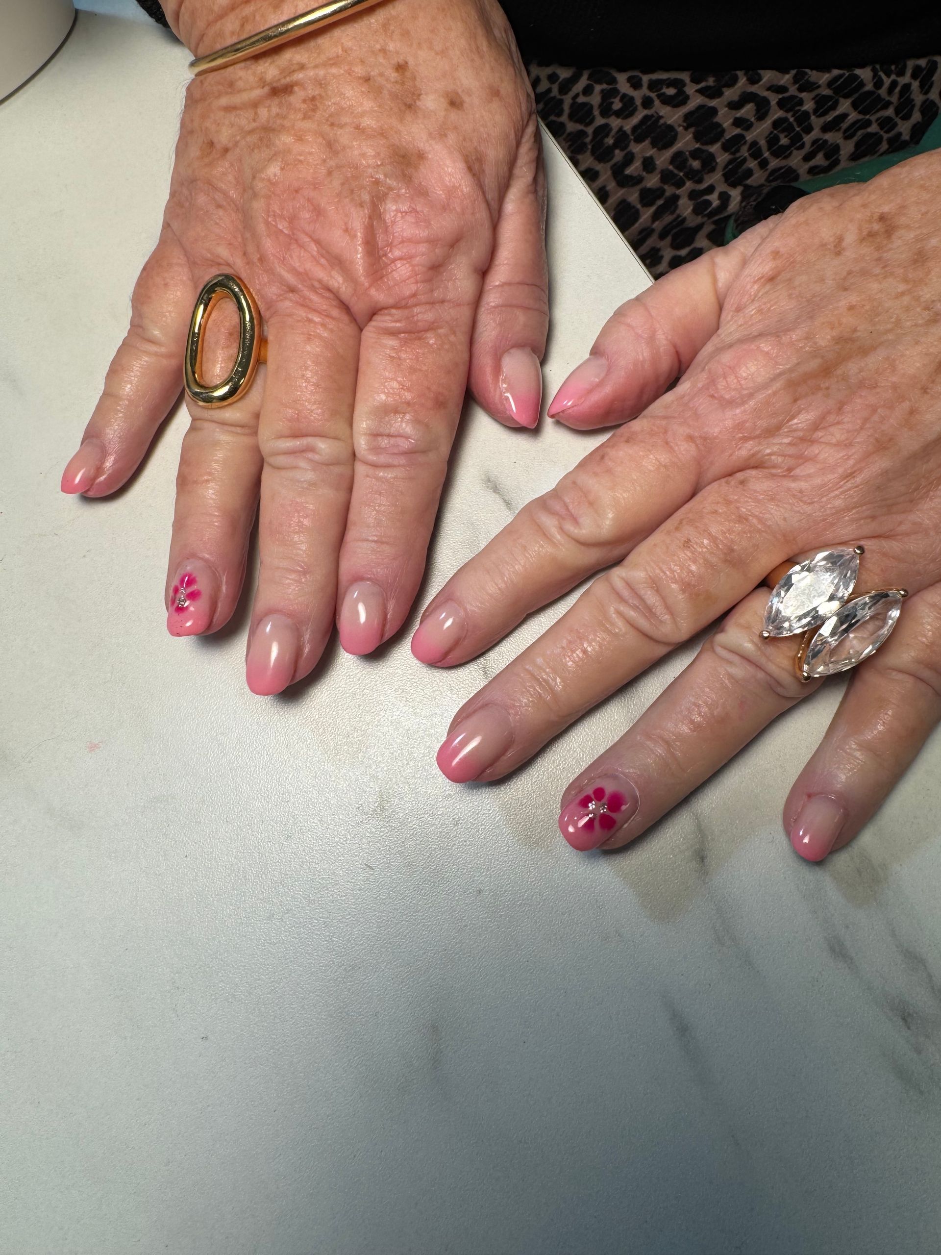 Deux mains aux ongles roses manucurés et ornées de grosses bagues reposent sur une surface blanche.