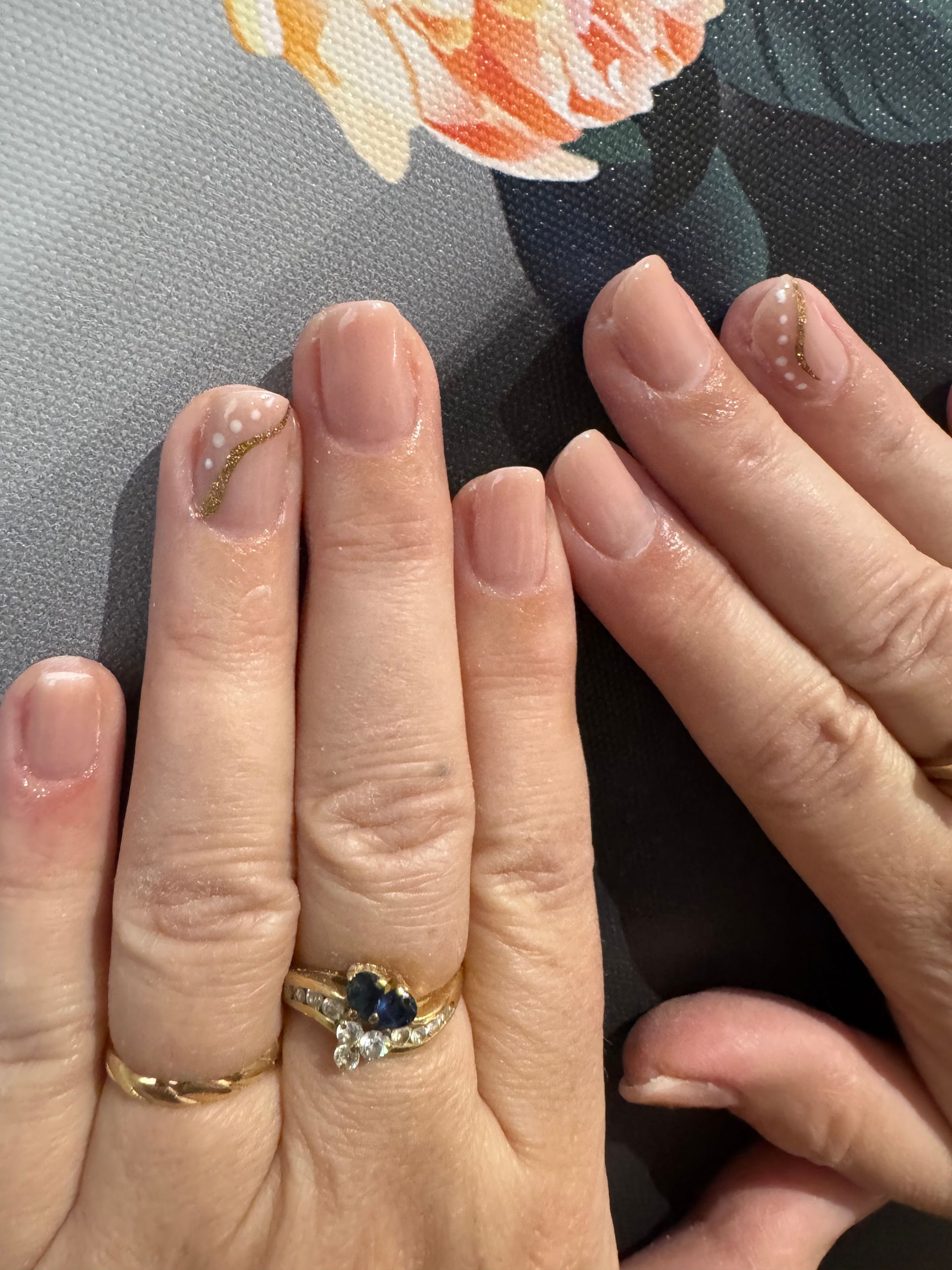 Des mains aux ongles nude et pailletés, ornées de bagues, sont posées près de fleurs orange sur une surface grise.