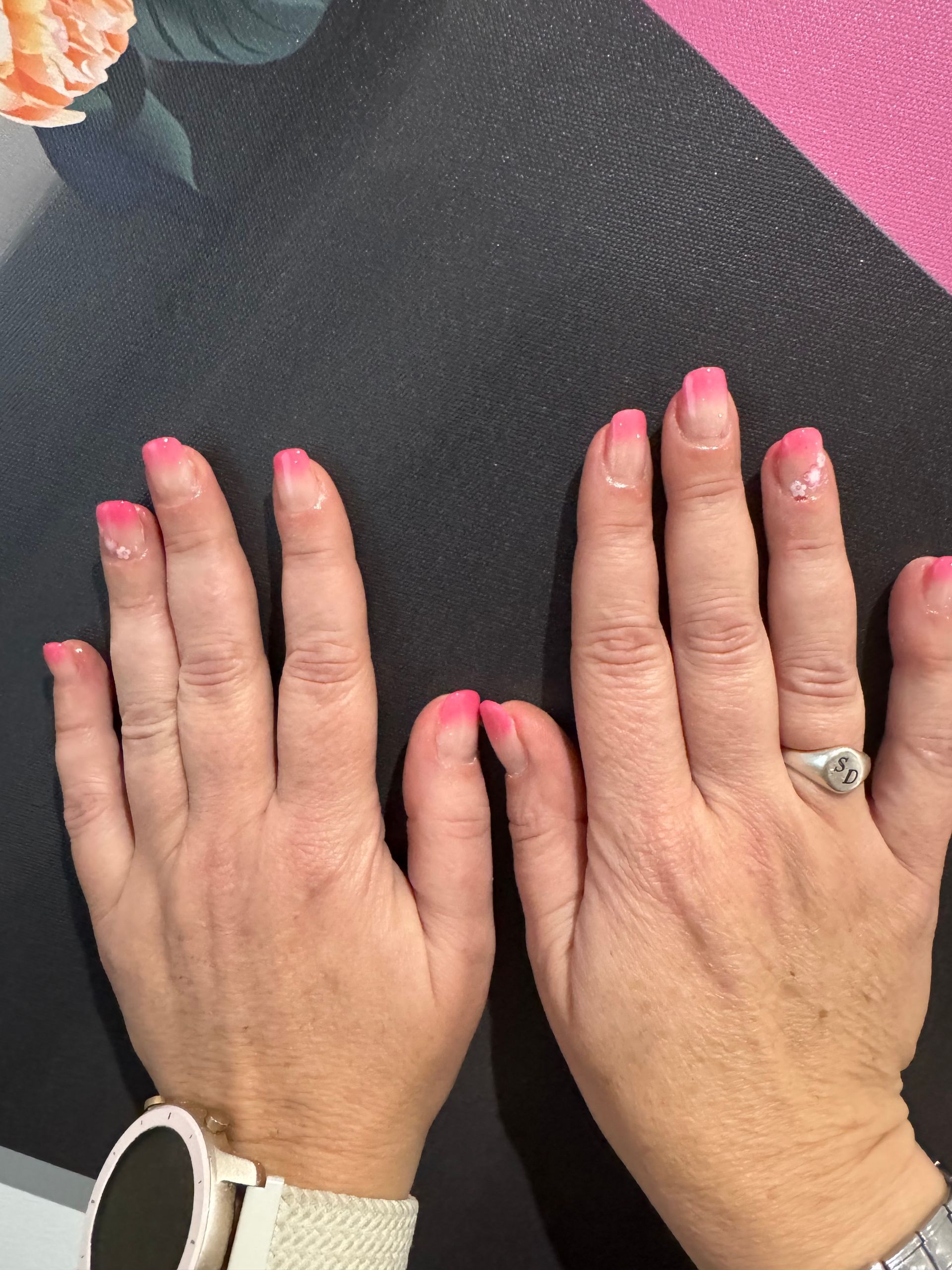 Deux mains aux ongles vernis de rose posées sur une surface noire, avec une bague argentée et une montre blanche visibles.
