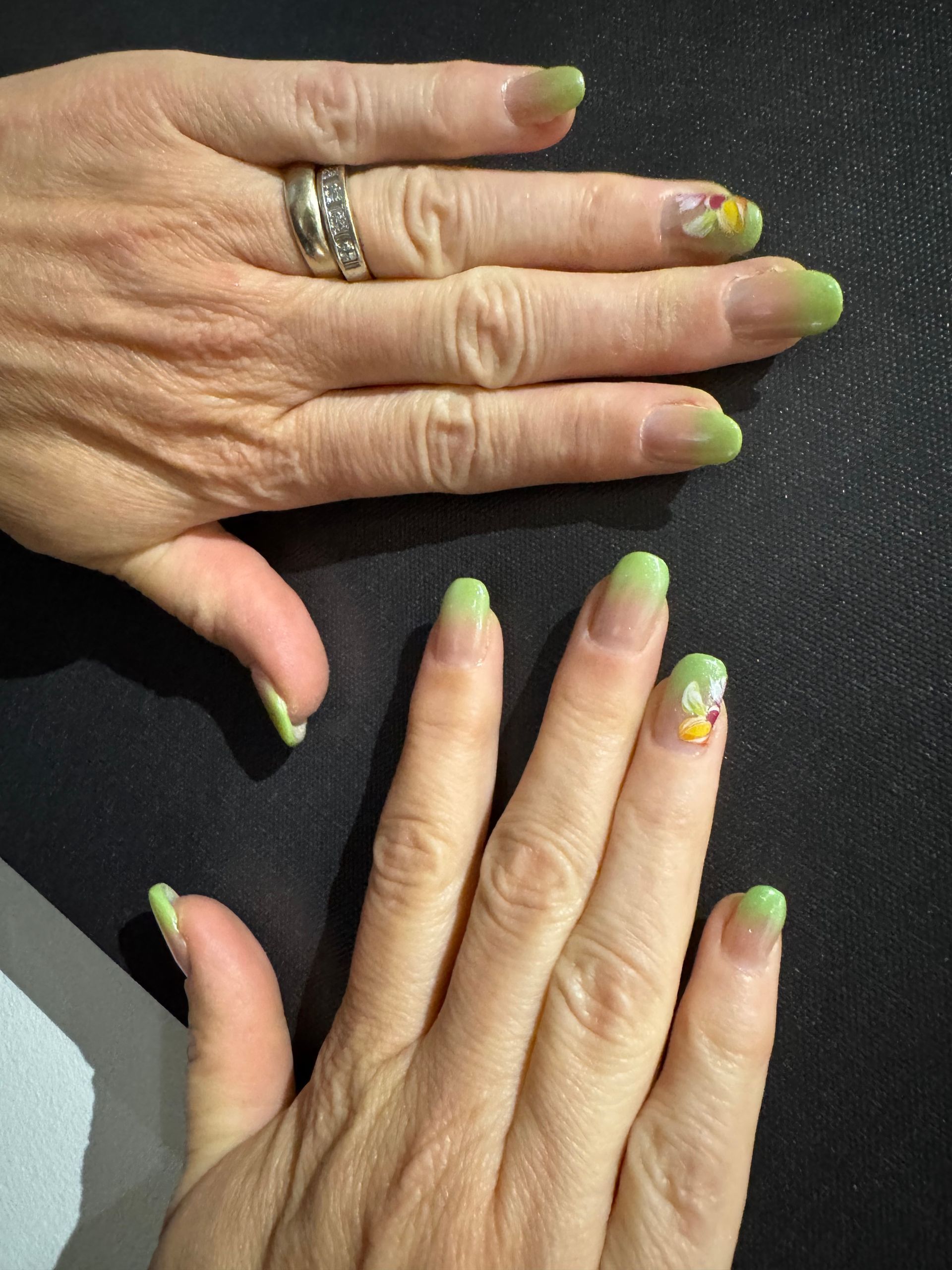 Deux mains aux ongles vernis vert fluo, l'une ornée d'une bague argentée, sur fond sombre.