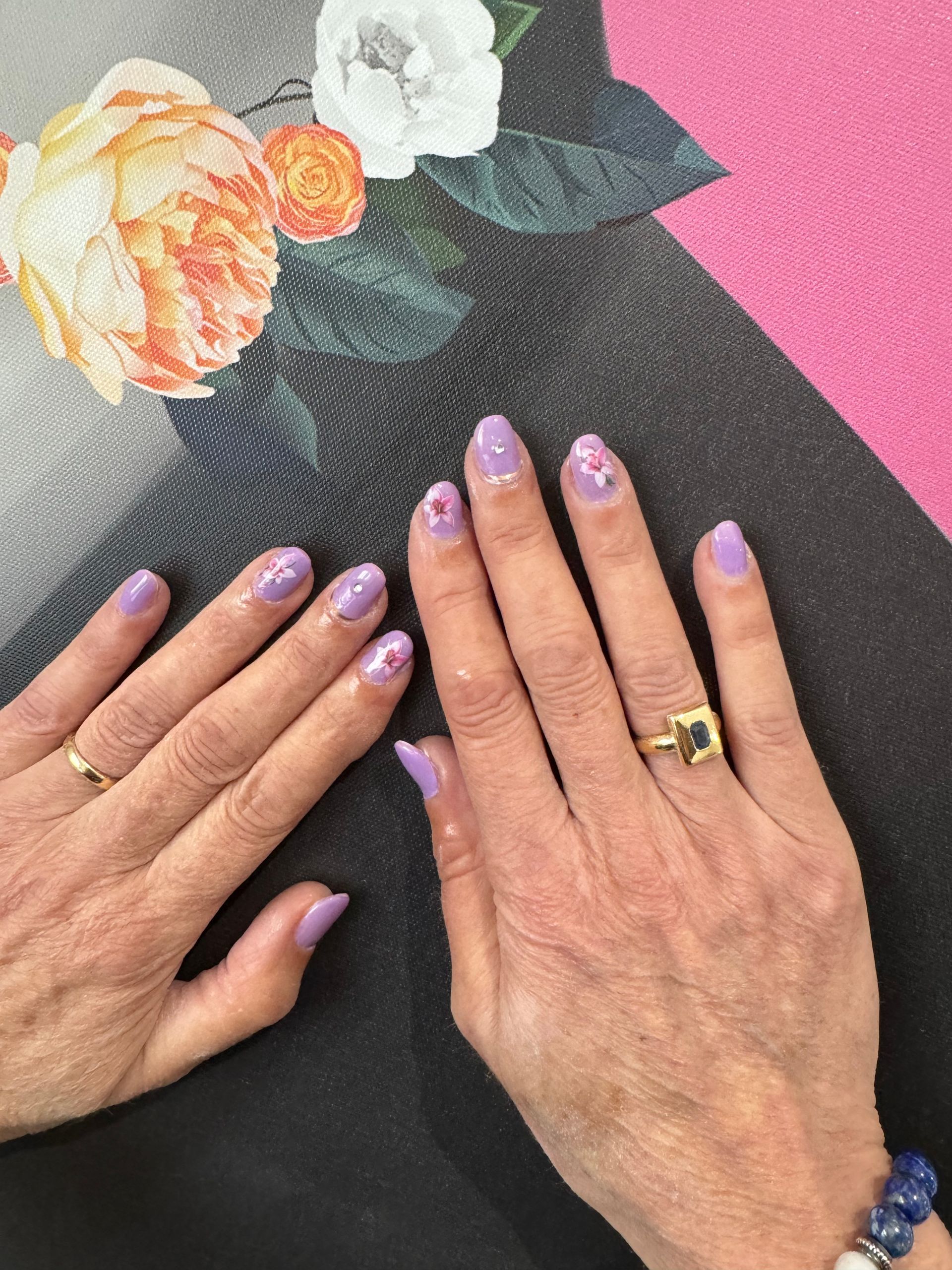 Deux mains aux ongles manucurés violets et aux bagues en or sur un fond floral et rose