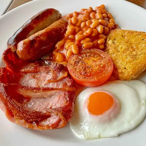 Un plato blanco cubierto con tocino, huevos, frijoles, tomates y salchichas.