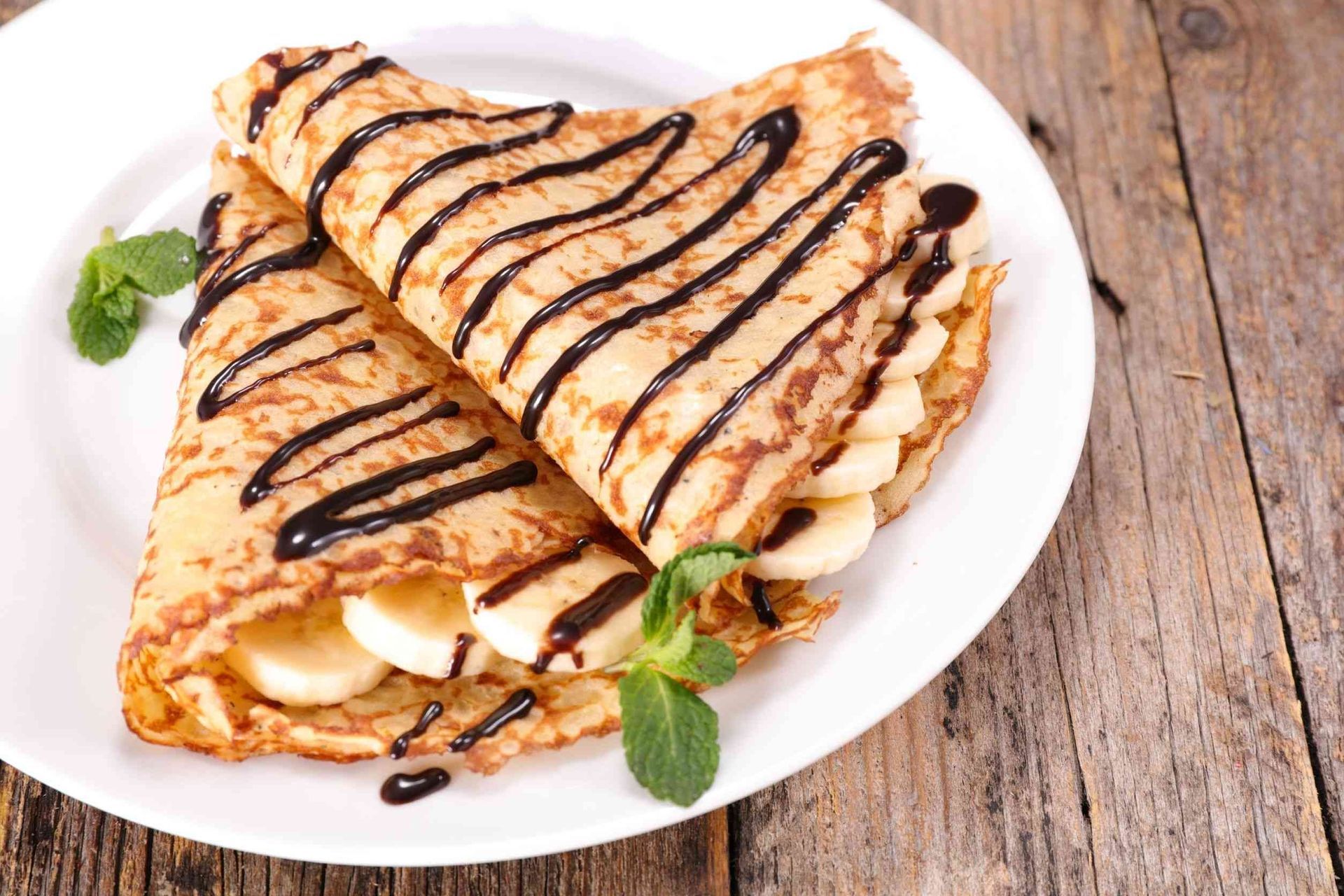 Dos panqueques con plátanos y salsa de chocolate en un plato blanco sobre una mesa de madera.