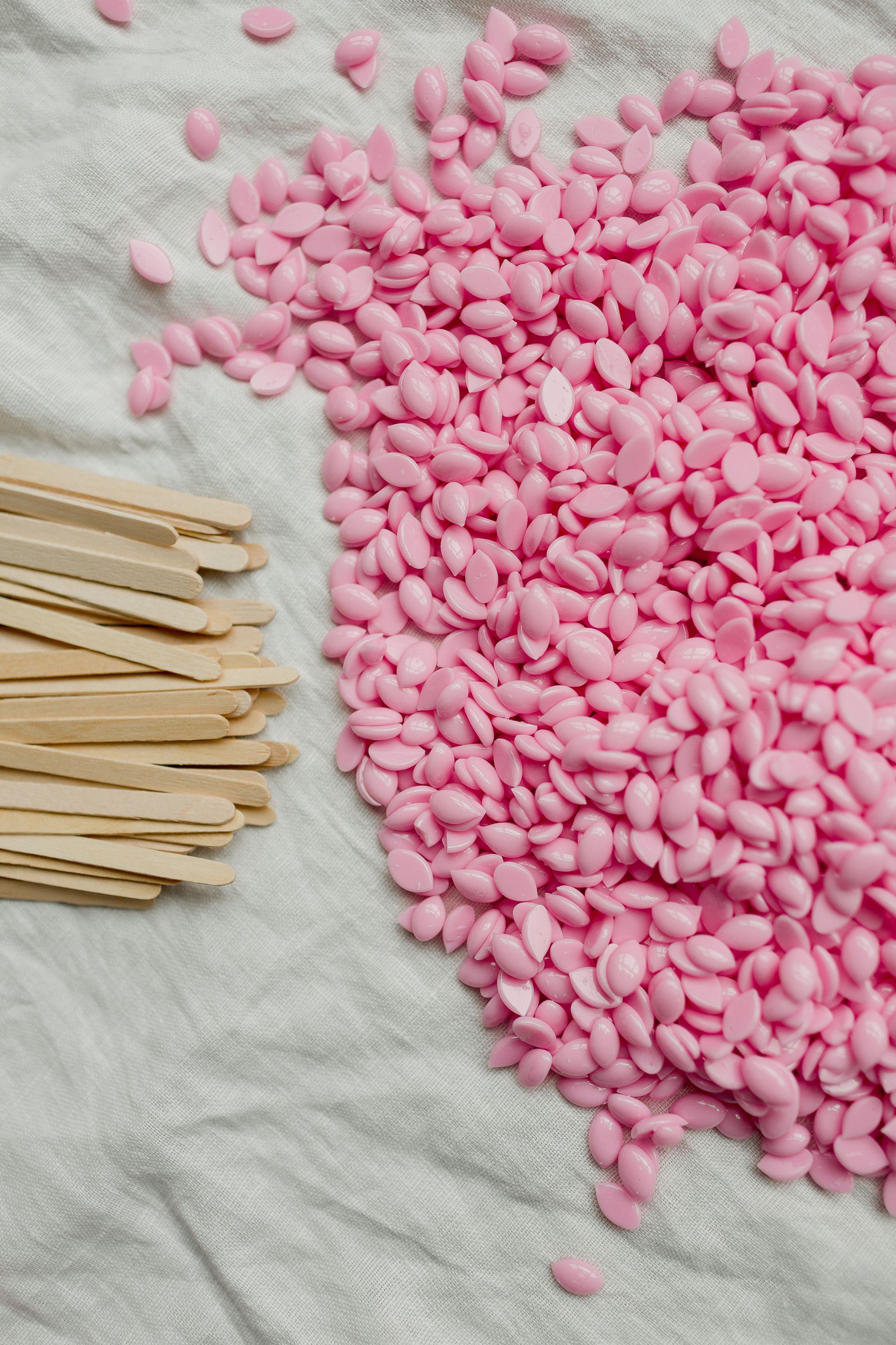 Perles de cire rose et applicateurs en bois sur une serviette blanche.